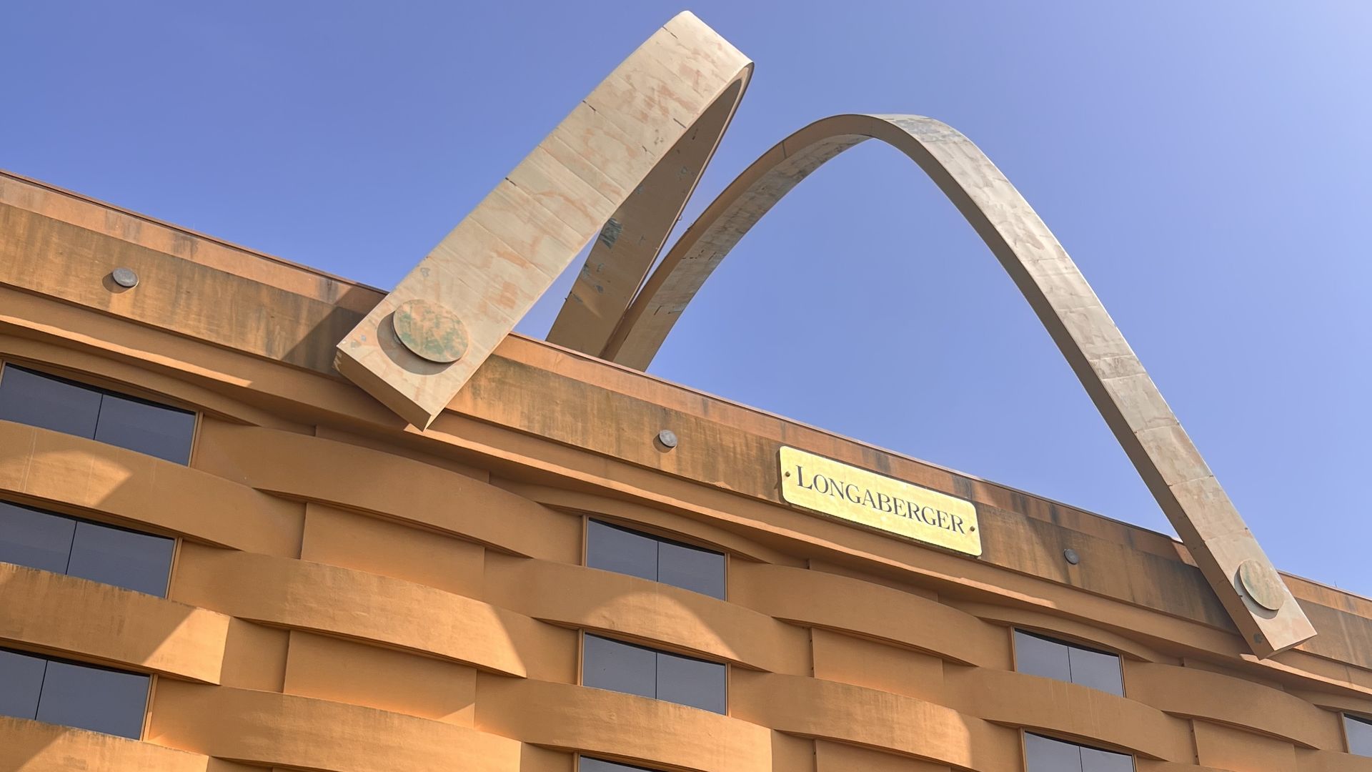 Orange building with a large metal arch sculpture on the roof; curved arches create a decorative top. A sign reads LONGABERGER, with a blue sky and green shrubs in front.