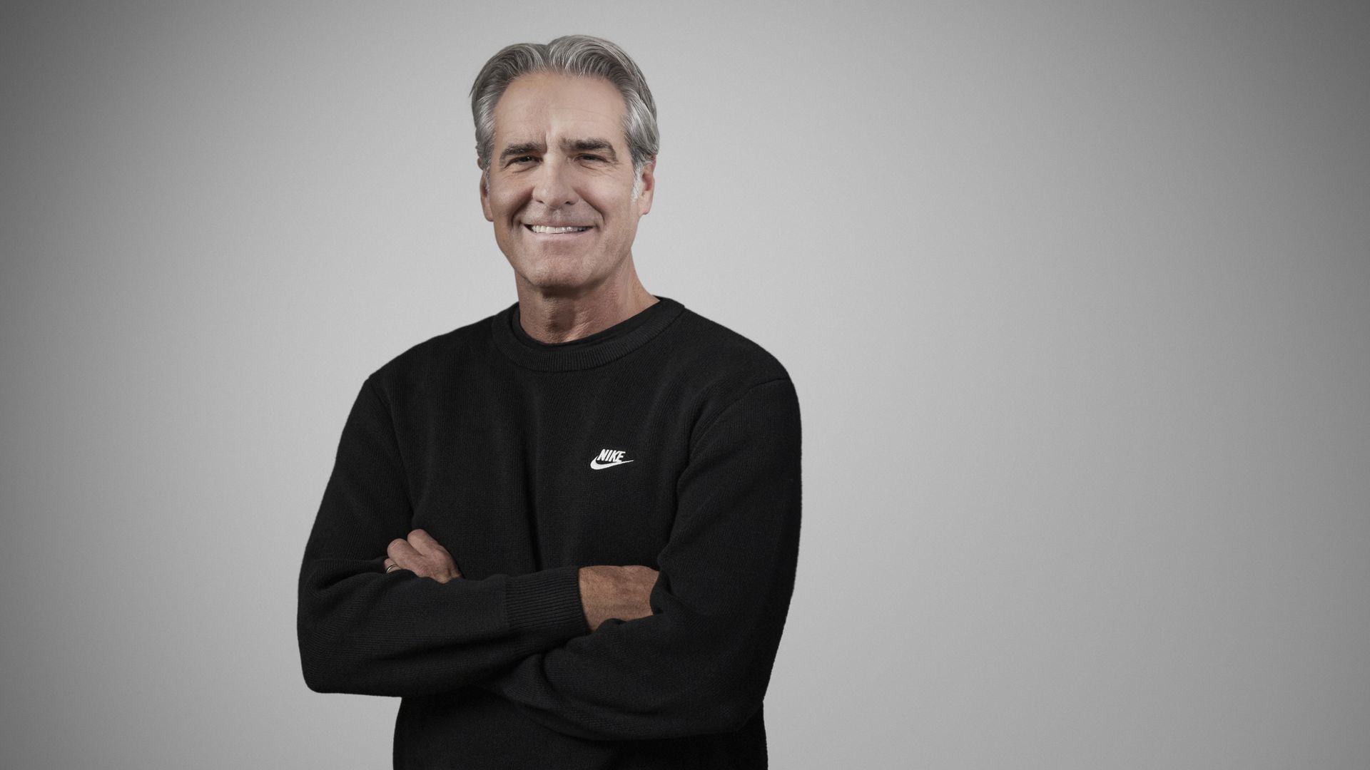 Middle-aged man with gray hair wearing a black Nike sweater stands with arms crossed, smiling against a plain gray background.