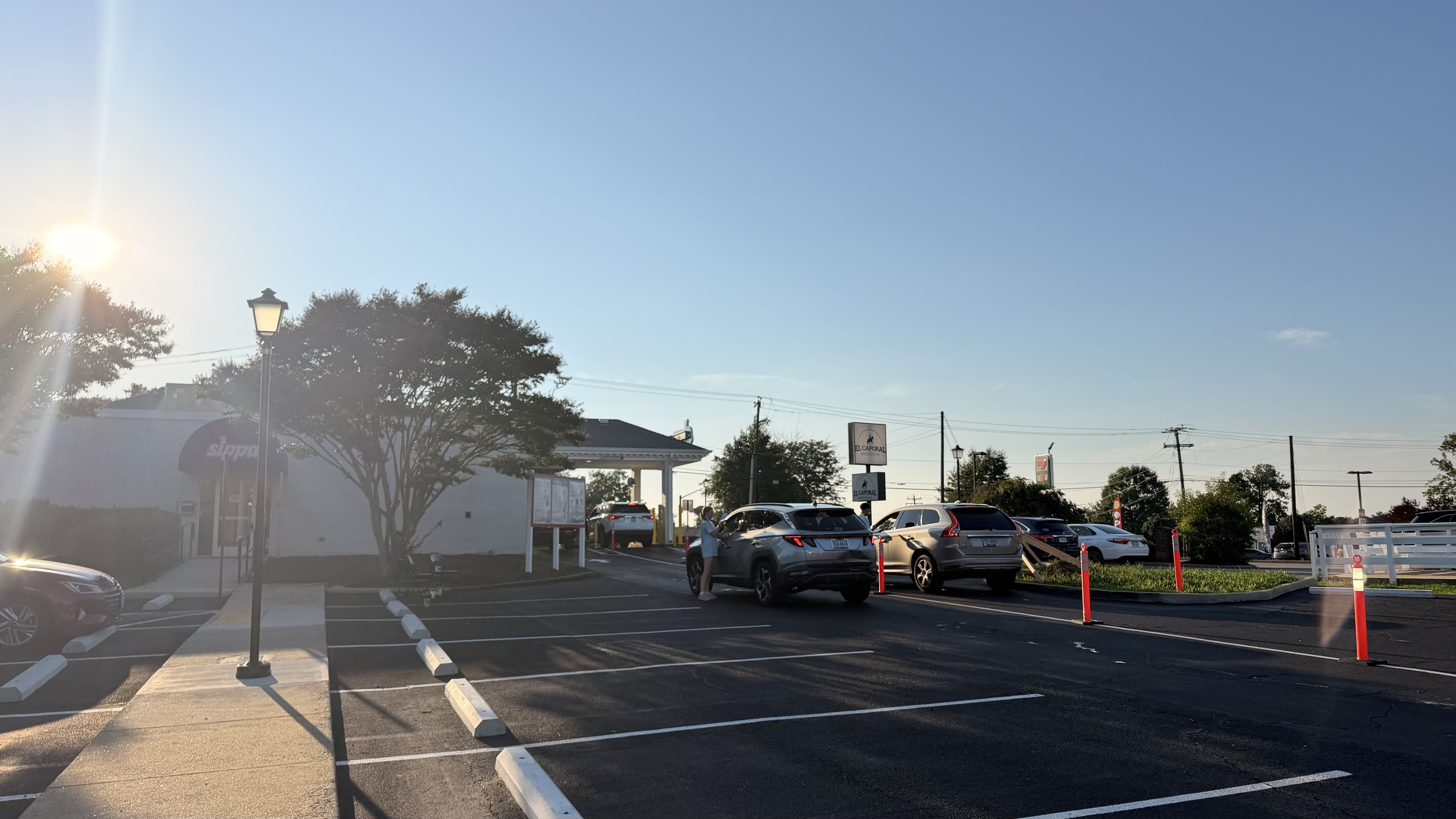 Sunny parking lot at a shopping center with lens flare from the left sun. Many cars parked, a lamppost, trees, and signs for "El Caporal." A person stands beside a car on the right.