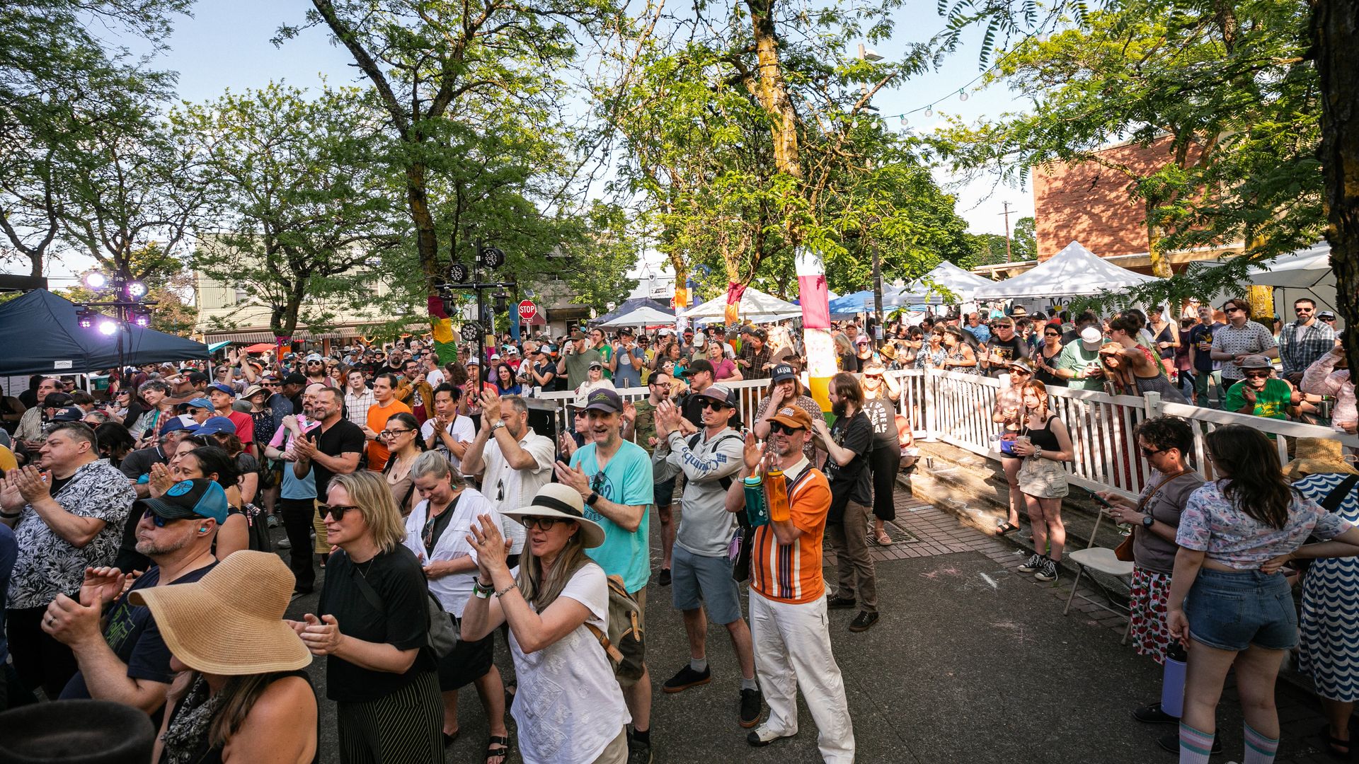 A group of people at an outdoor festival clap their hands near a performance stage.