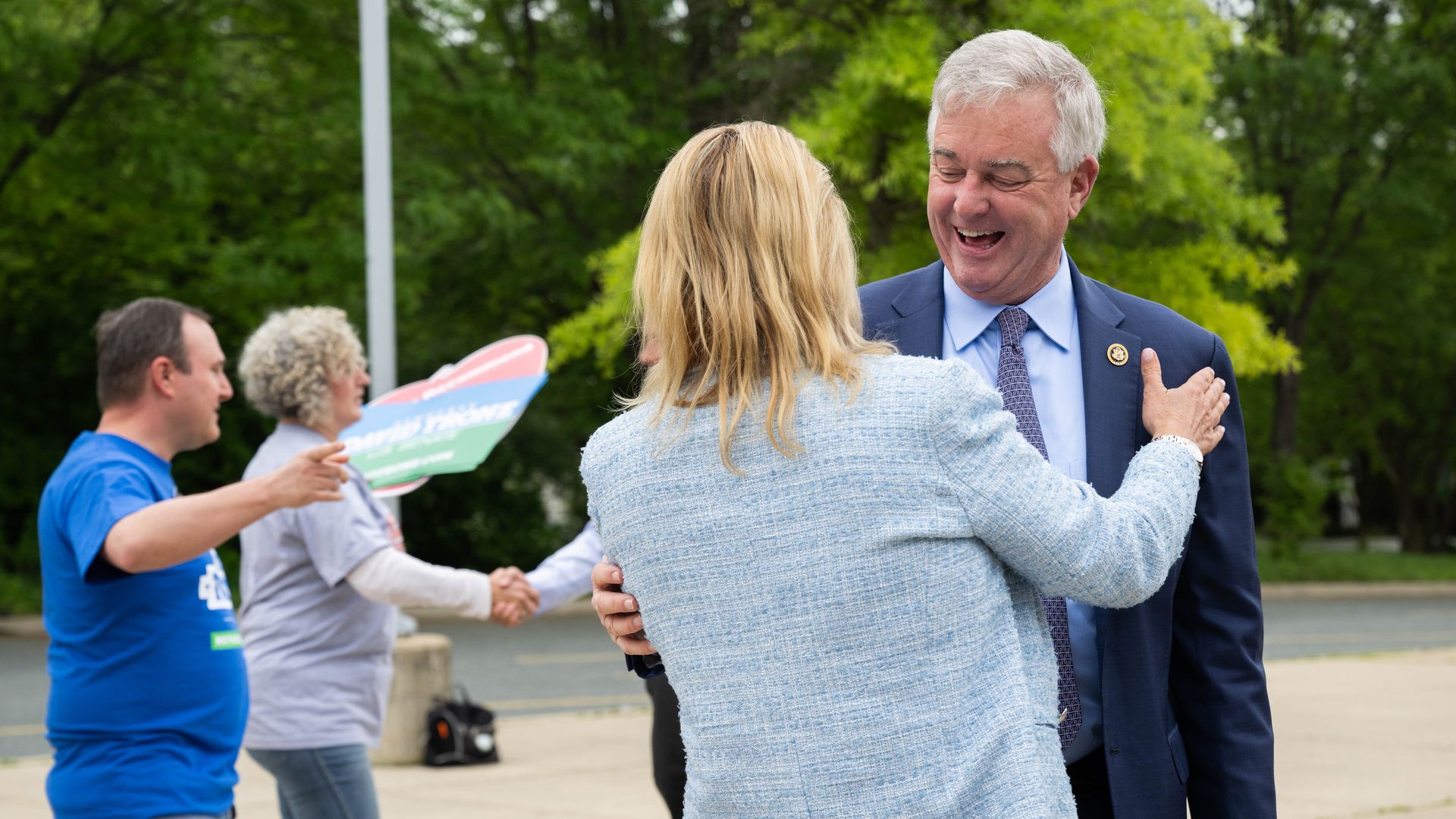 An older man in a navy suit and lavender tie smiles as a blonde woman in a light blue blazer embraces him outdoors; in the background, two people shake hands near a colorful sign.