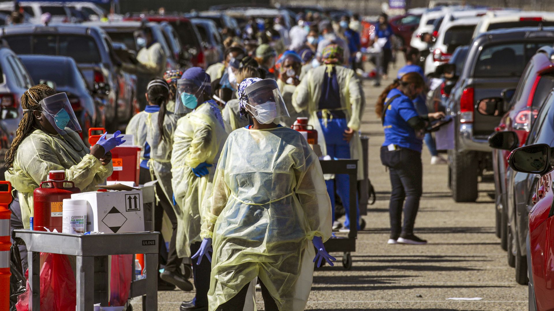 People in PPE and medical gear administer vaccines with a long line of cars behind them