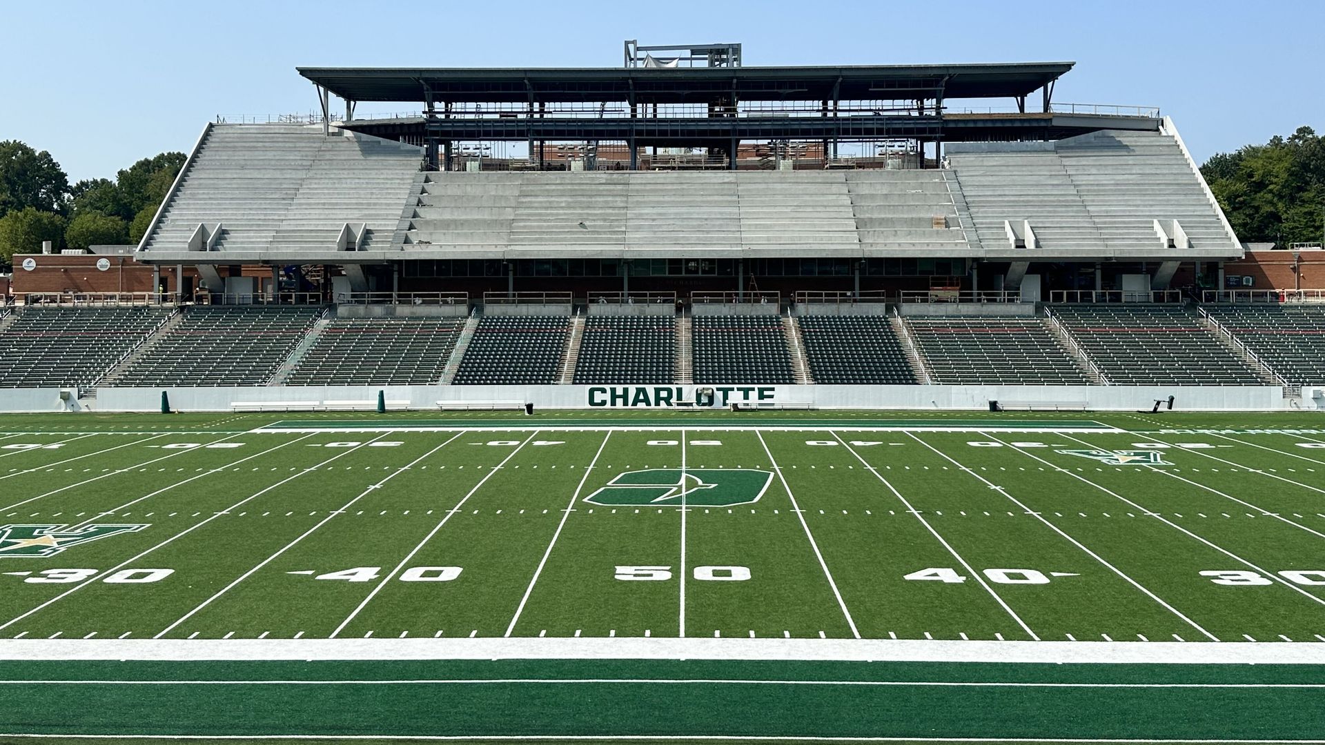 Wide view of an empty outdoor football stadium with a green field, white yard lines, and CHARLOTTE on the front wall. Concrete stands and a steel press box sit beneath a clear blue sky.