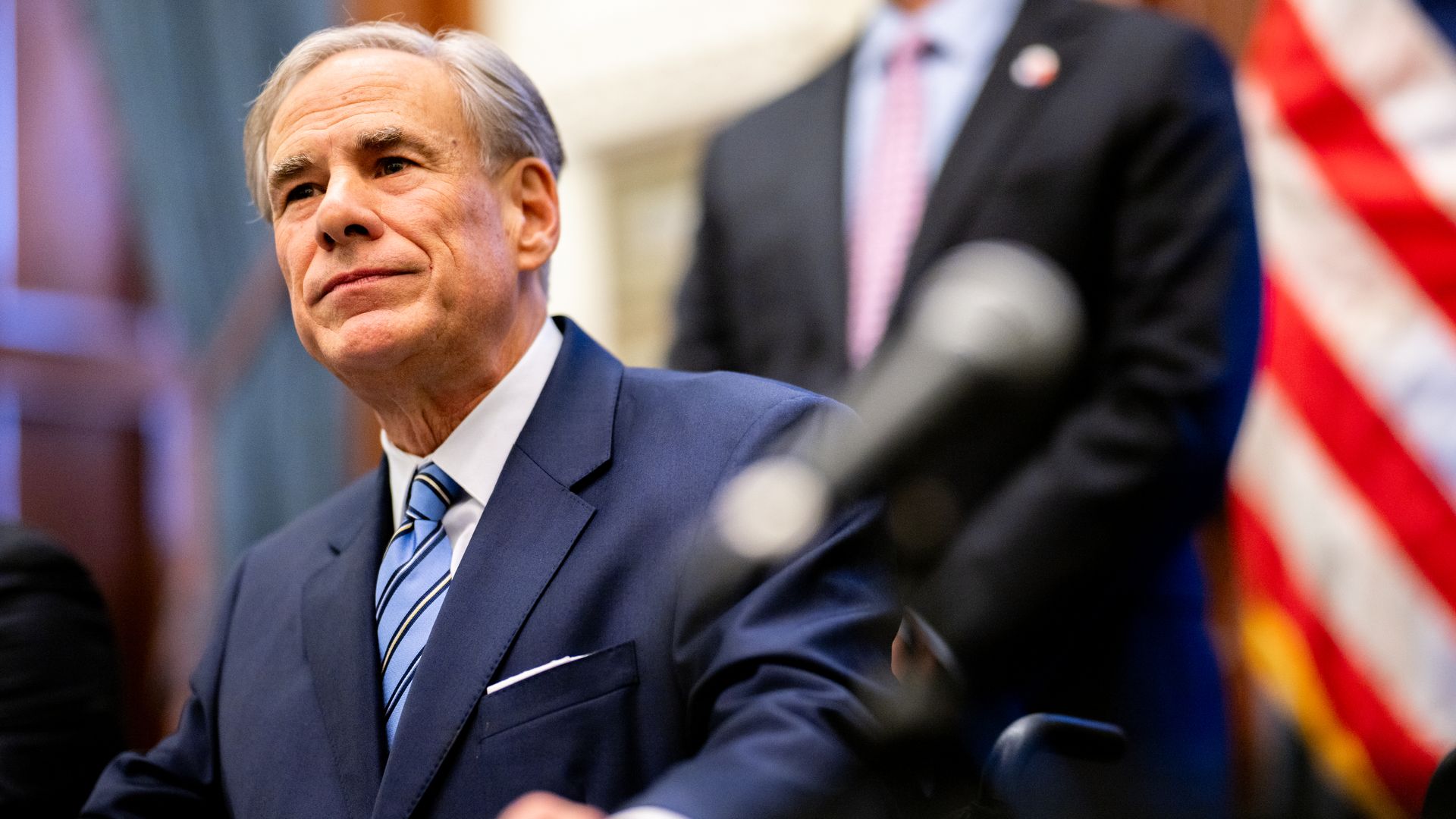 Gov. Greg Abbott is seen with a blurred American flag in the background and a blurred microphone in front of him.