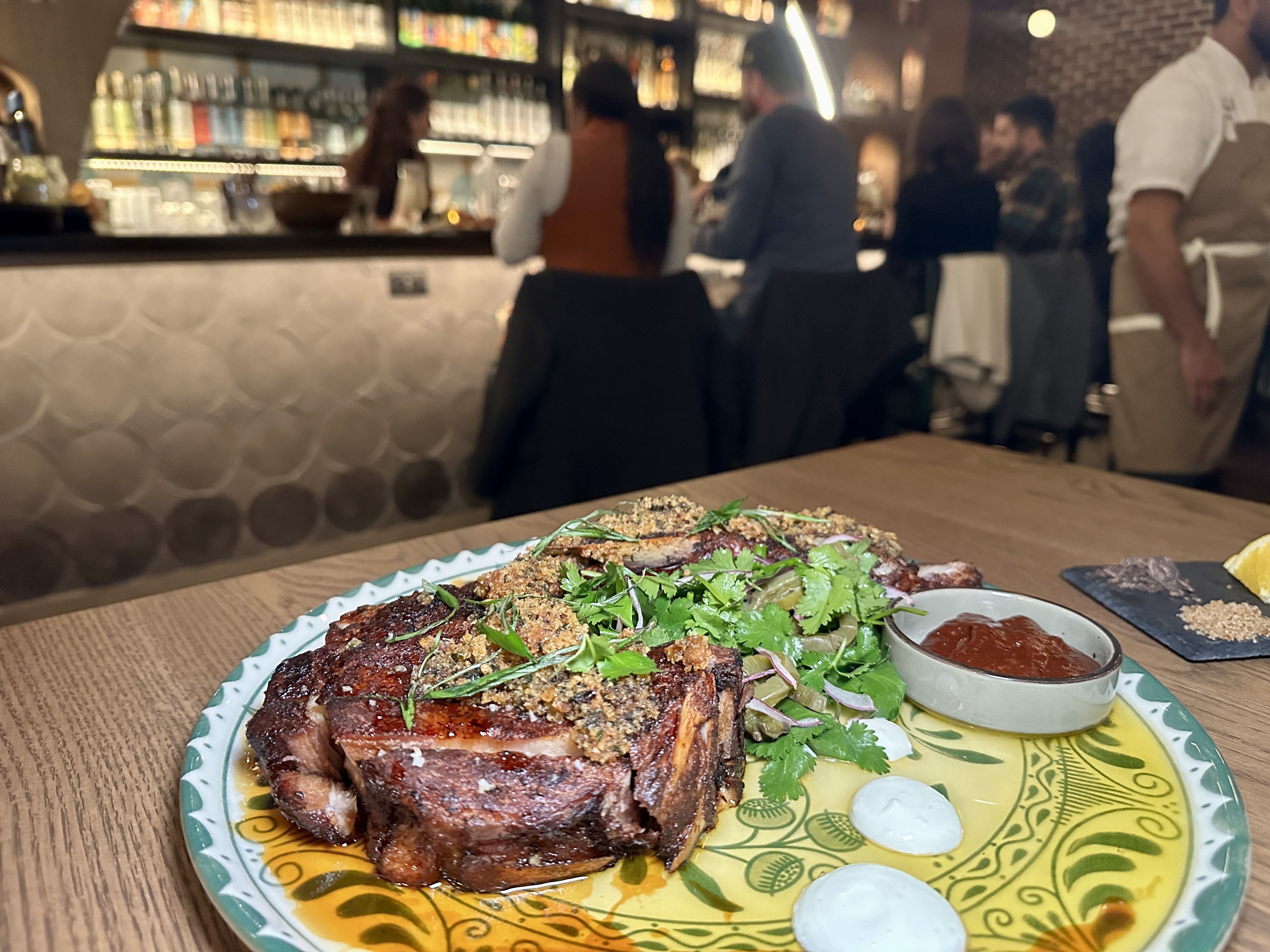 Thick roasted meat topped with breadcrumbs and fresh herbs on a colorful ceramic plate, with greens and onions; a small dish of red sauce beside. Blurred bar patrons and staff in the background.