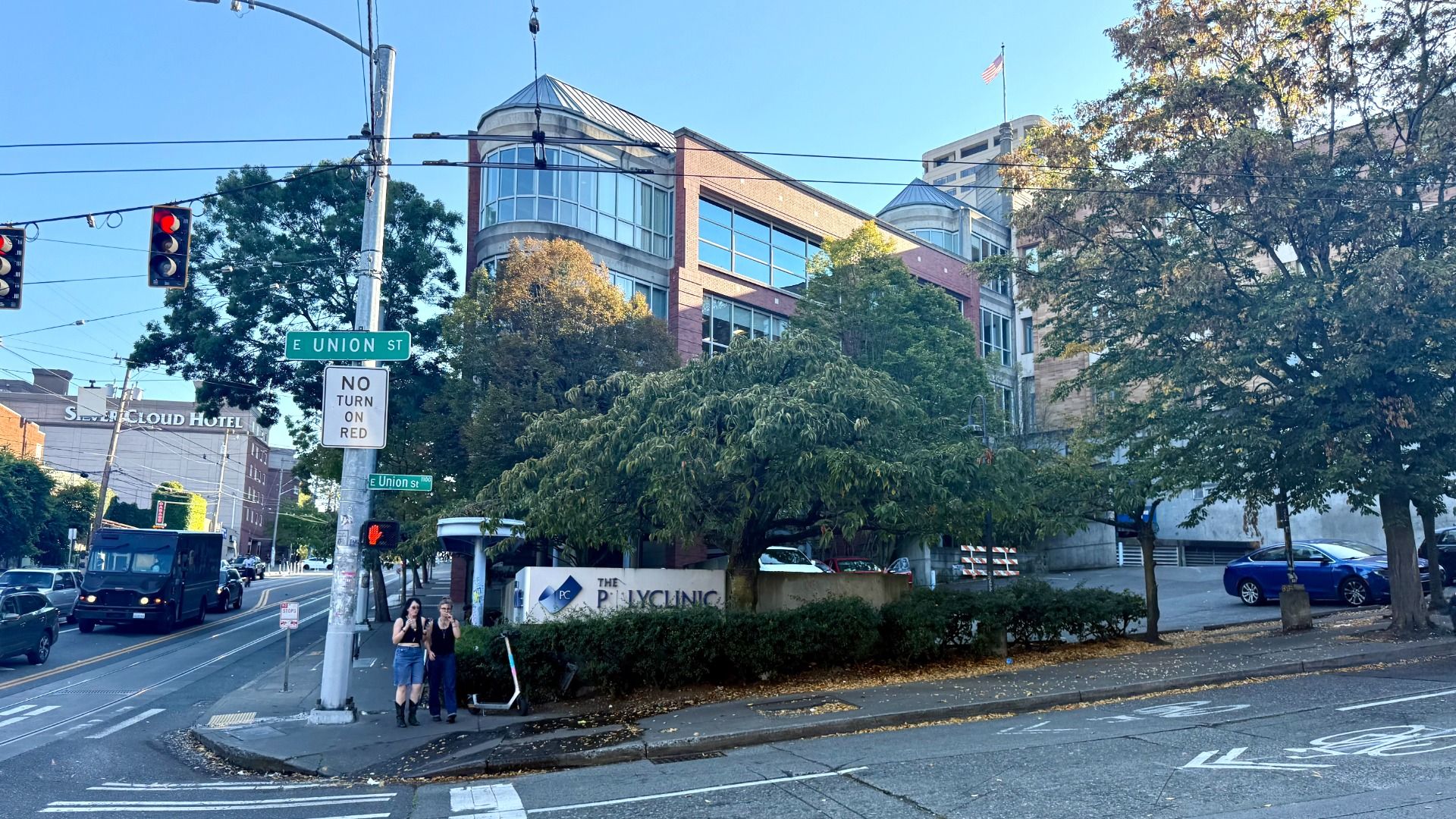 Intersection of E Union St with traffic lights, street signs, trees, and people standing on the sidewalk. Red brick building with glass windows and a sign for "The Polyclinic."