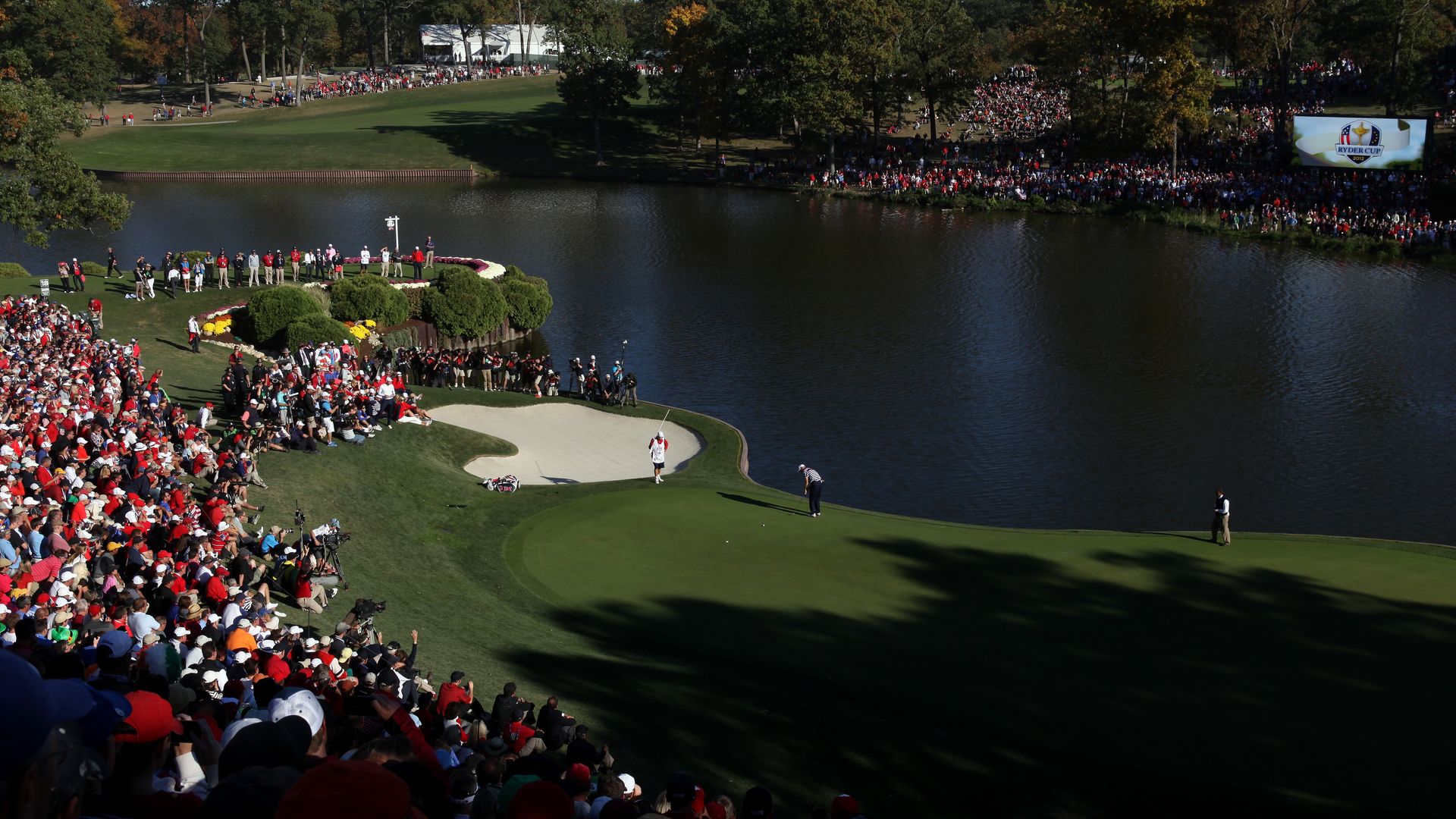 Panoramic view of a crowded golf tournament by a lake, with fans along the fairways, autumn trees in the background, and a large screen displaying Ryder Cup 2012 on the right.