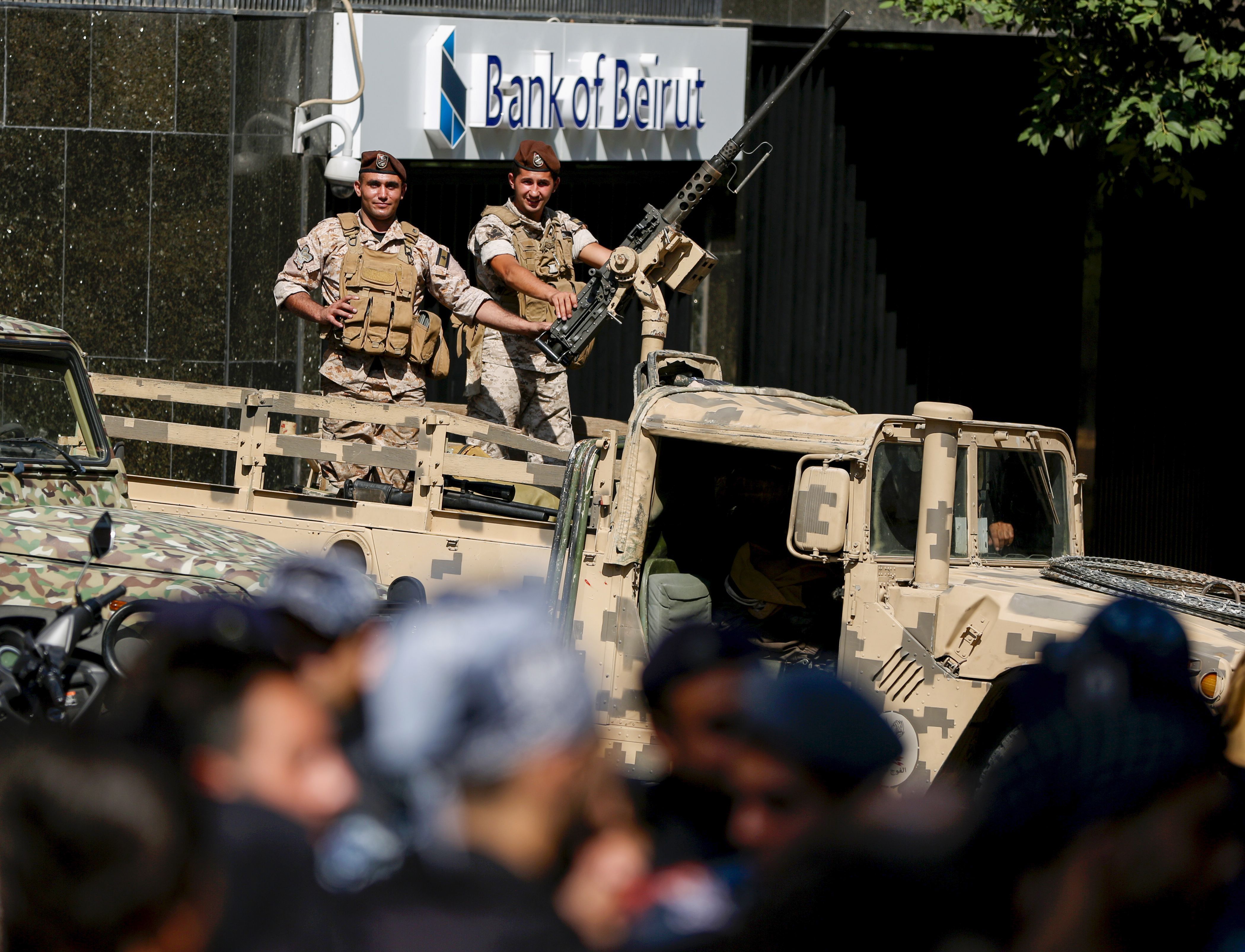 In this image, two soldiers holding a gun on top of an armored truck watch protestors from a distance. The truck is parked in front of a bank