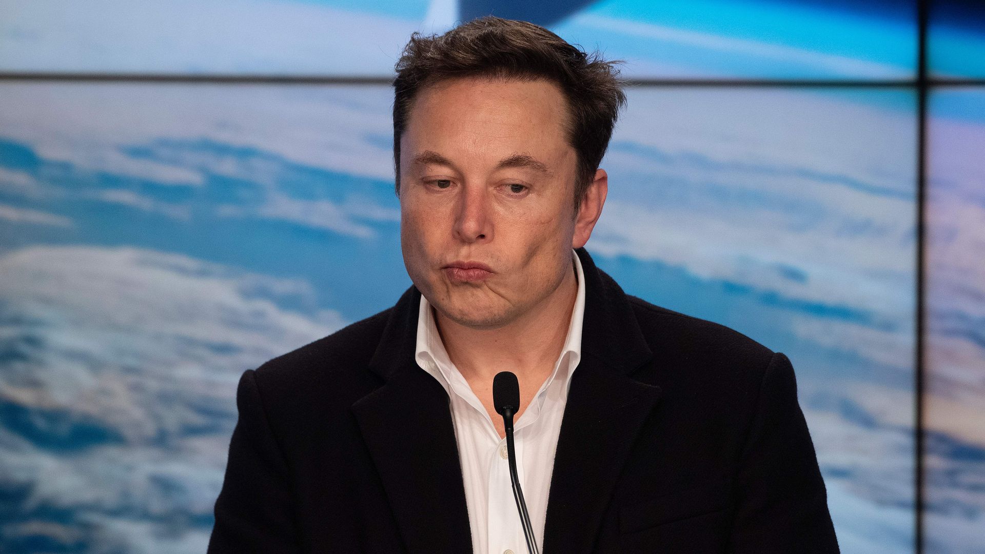 In this image, Elon Musk looks down while standing in front of a microphone. He's wearing a suit with no tie.