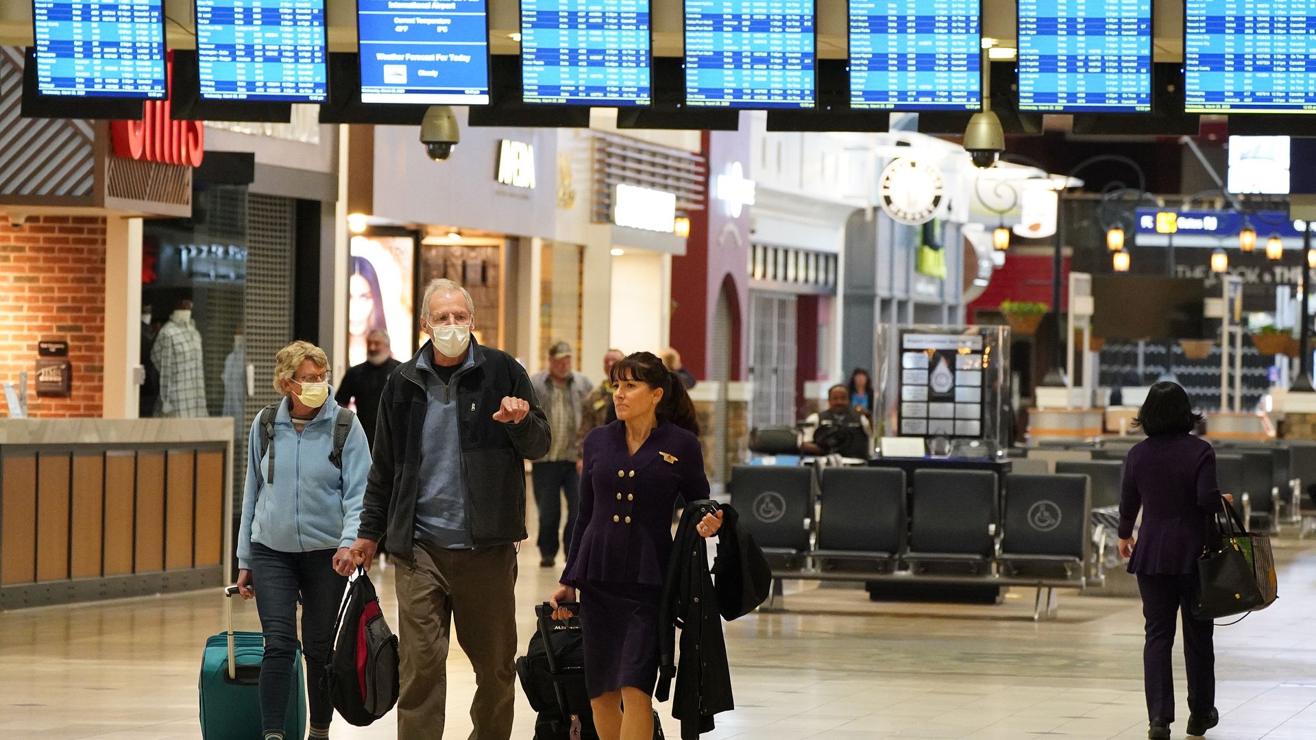 In this image, two people wearing face masks walk through an airport while a flight attendant walks next to them