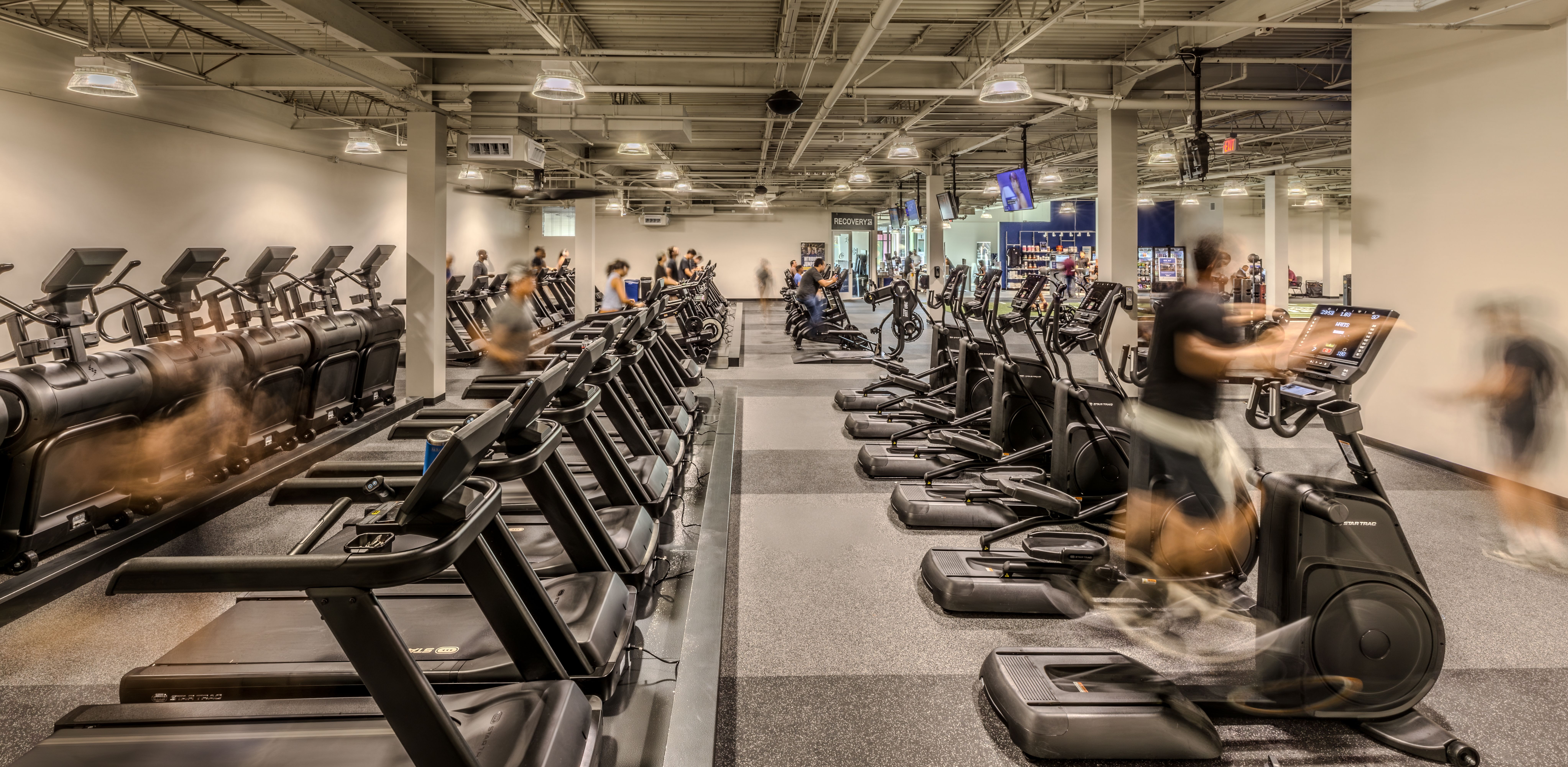 Spacious gym with row of treadmills on the left and ellipticals on the right; motion-blurred exercisers, exposed ceiling pipes, bright lights, and a recovery area with vending machines in back.