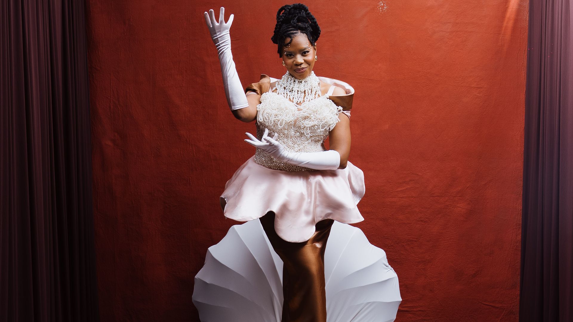 Perrine Deshield Met Gala Charlotte. Woman in a sparkling white beaded gown and long white gloves stands on a round white pedestal, waving. She poses before a red backdrop with maroon curtains in a studio setting.