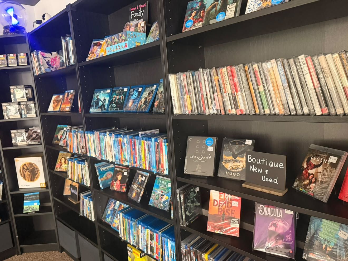 Black shelves filled with DVD and Blu-ray cases in a store. A chalkboard sign reads "Boutique New & Used" as colorful movie titles line the racks, with a blue light visible on the left.
