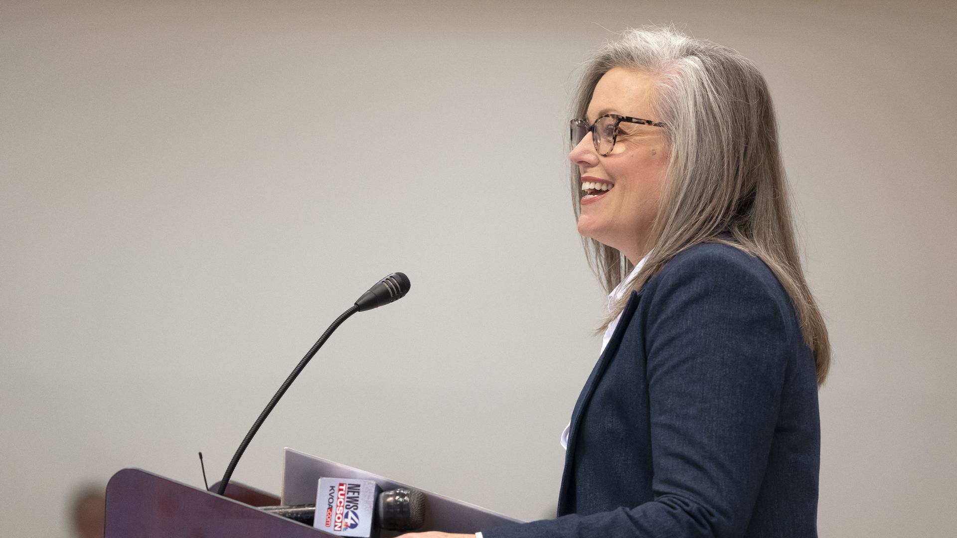 A smiling woman wearing glasses and a blue jacket is pictured from the side at a lectern with a microphone. 