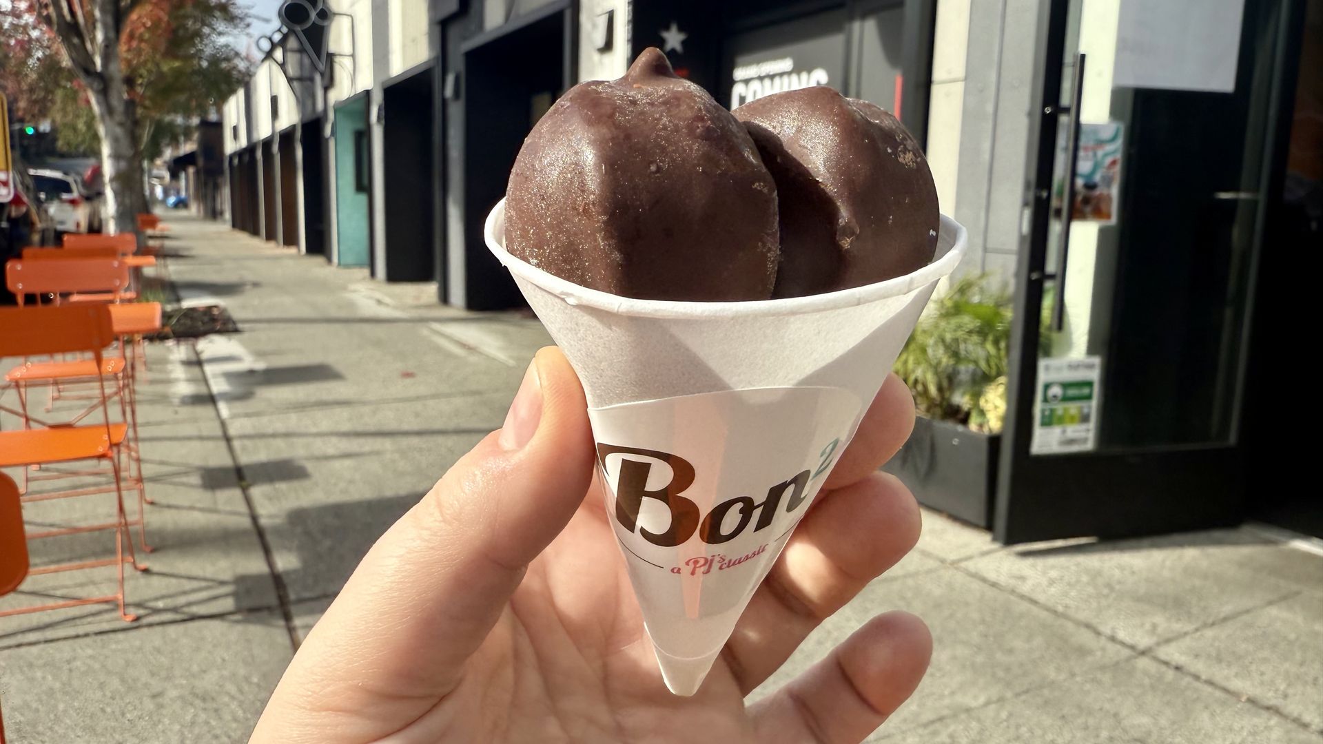 Hand holding a white paper cone with two chocolate-covered ice cream scoops outside a storefront on a sunny urban sidewalk with orange chairs and autumn trees.