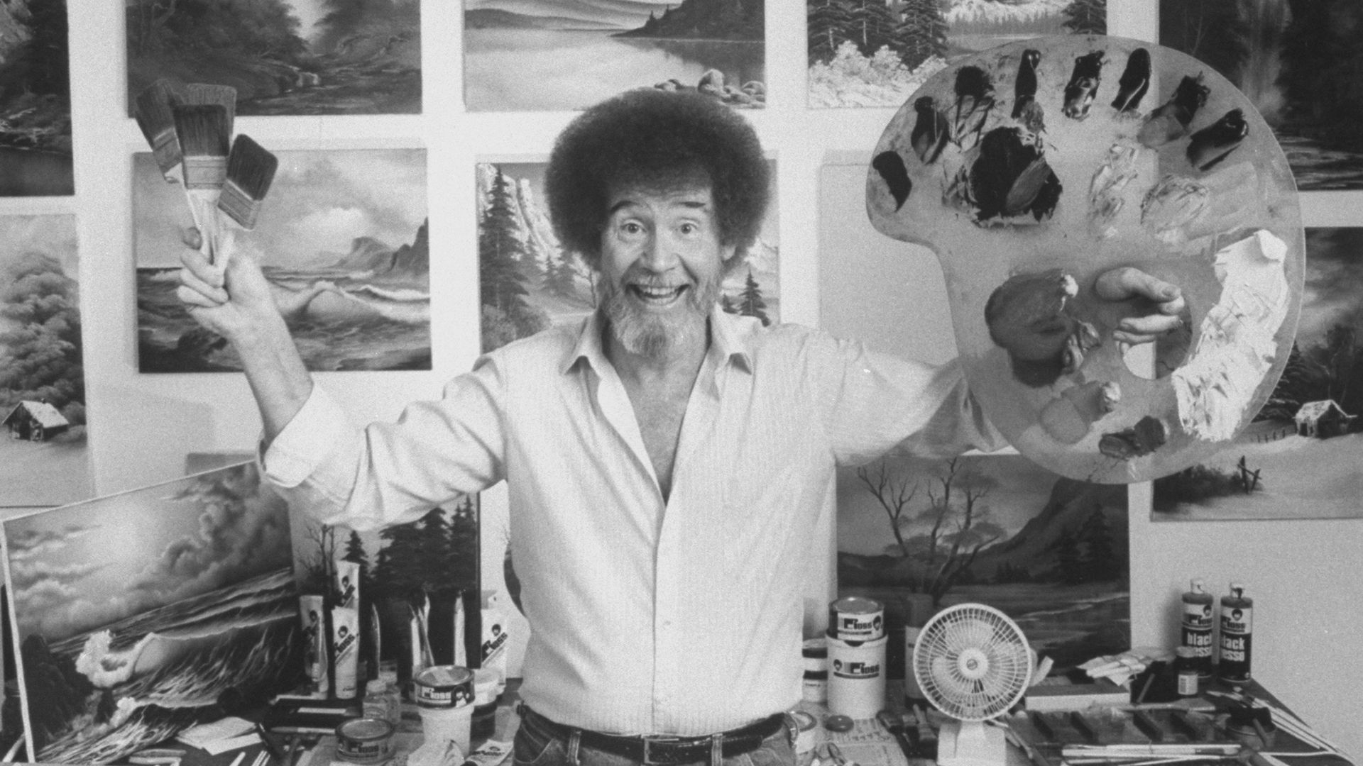 A painter holds a palette and brush with landscape paintings on the wall behind them