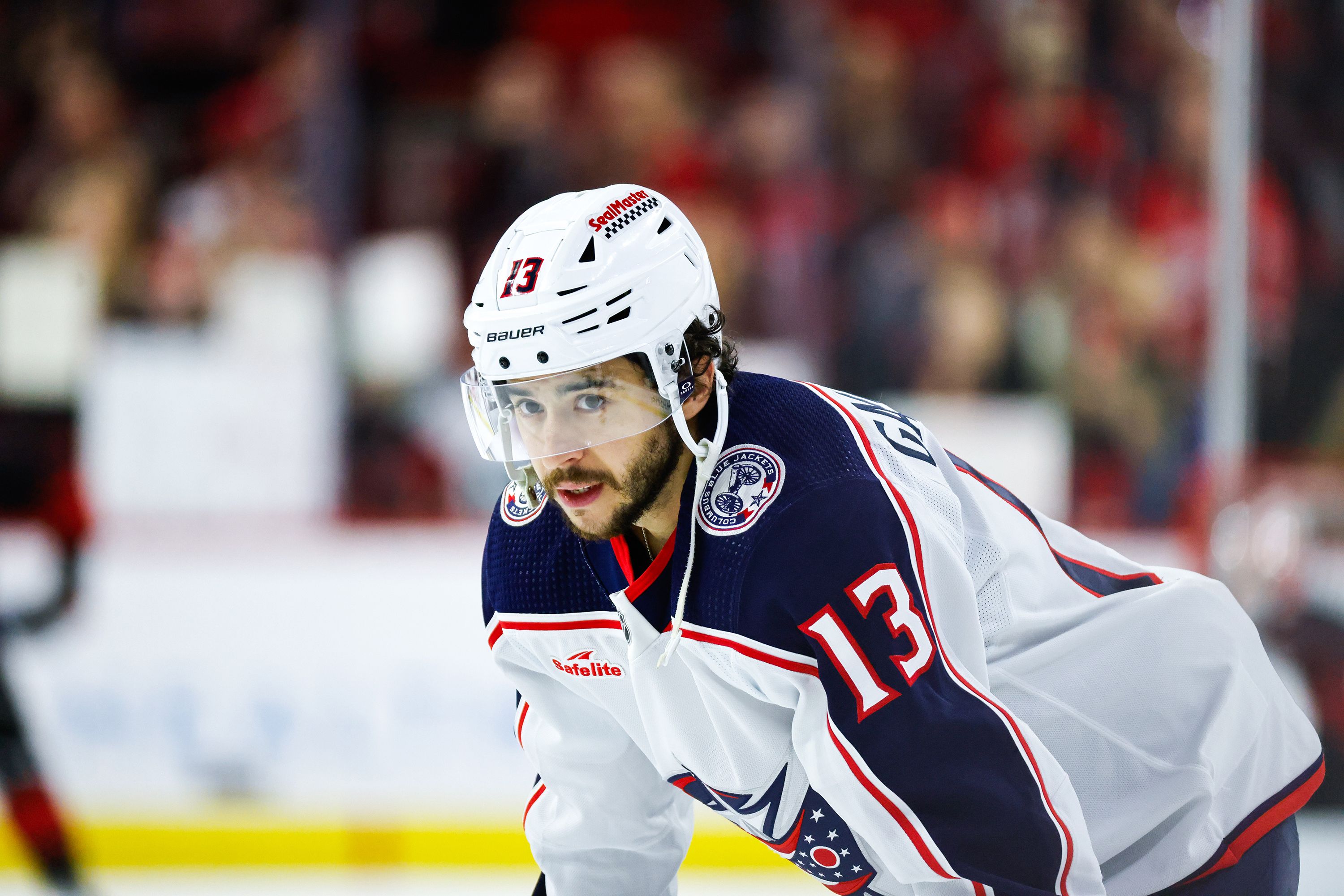 A hockey player in a white helmet and jersey bends over on his stick during a break in a game