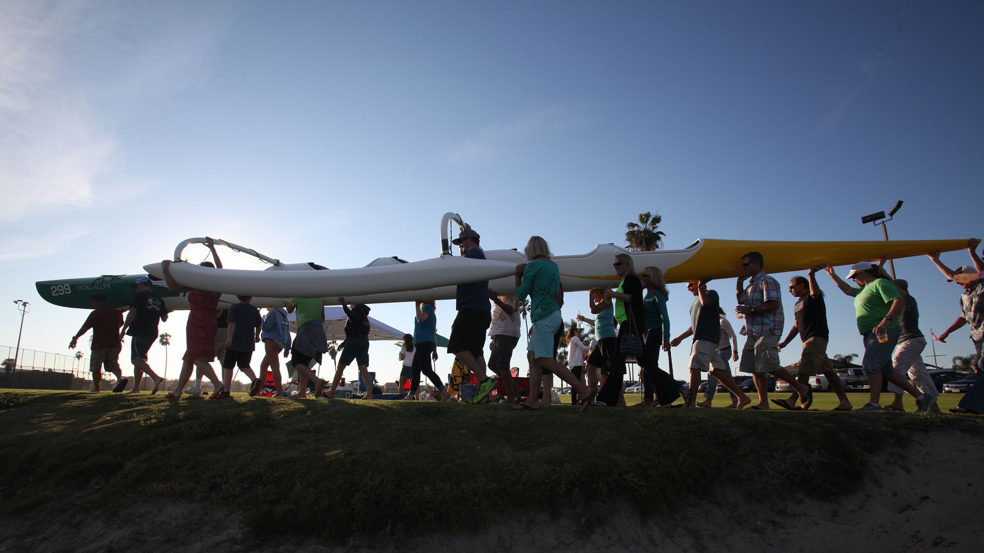 Group of people walks along a grassy dune, carrying a long white and yellow watercraft above their heads; clear blue sky, a palm tree in the distance, and onlookers nearby.