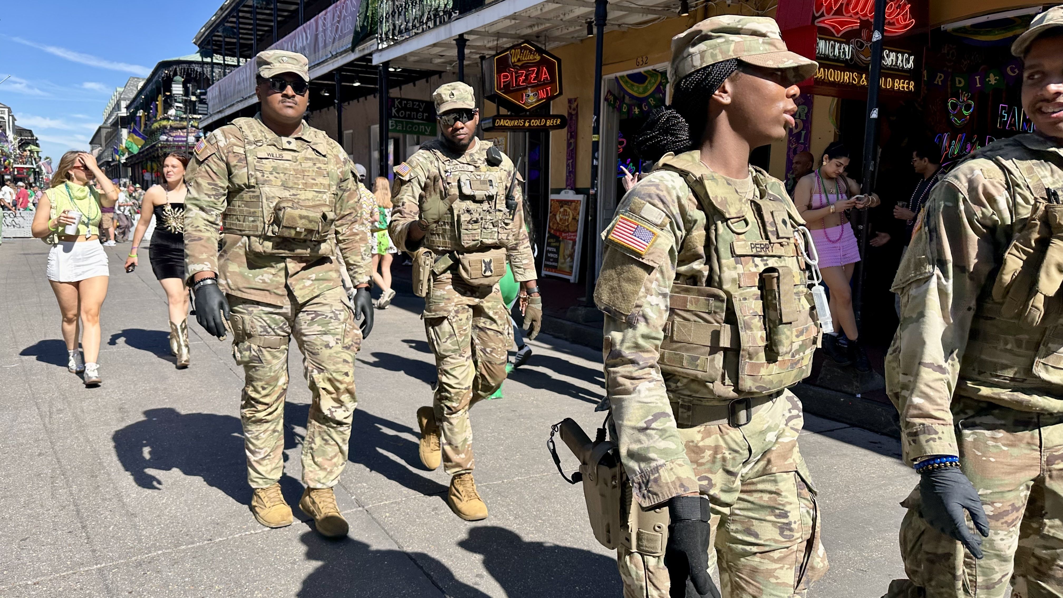 Four Louisiana National Guard soldiers in camouflage uniforms walking on a sunny street with people in casual clothing and neon signs for Willie's Pizza and drinks in the background.