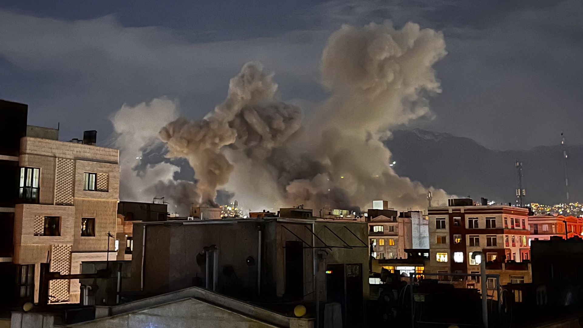 Nighttime cityscape with multiple large plumes of thick gray smoke rising behind buildings under a dark sky with faint clouds.
