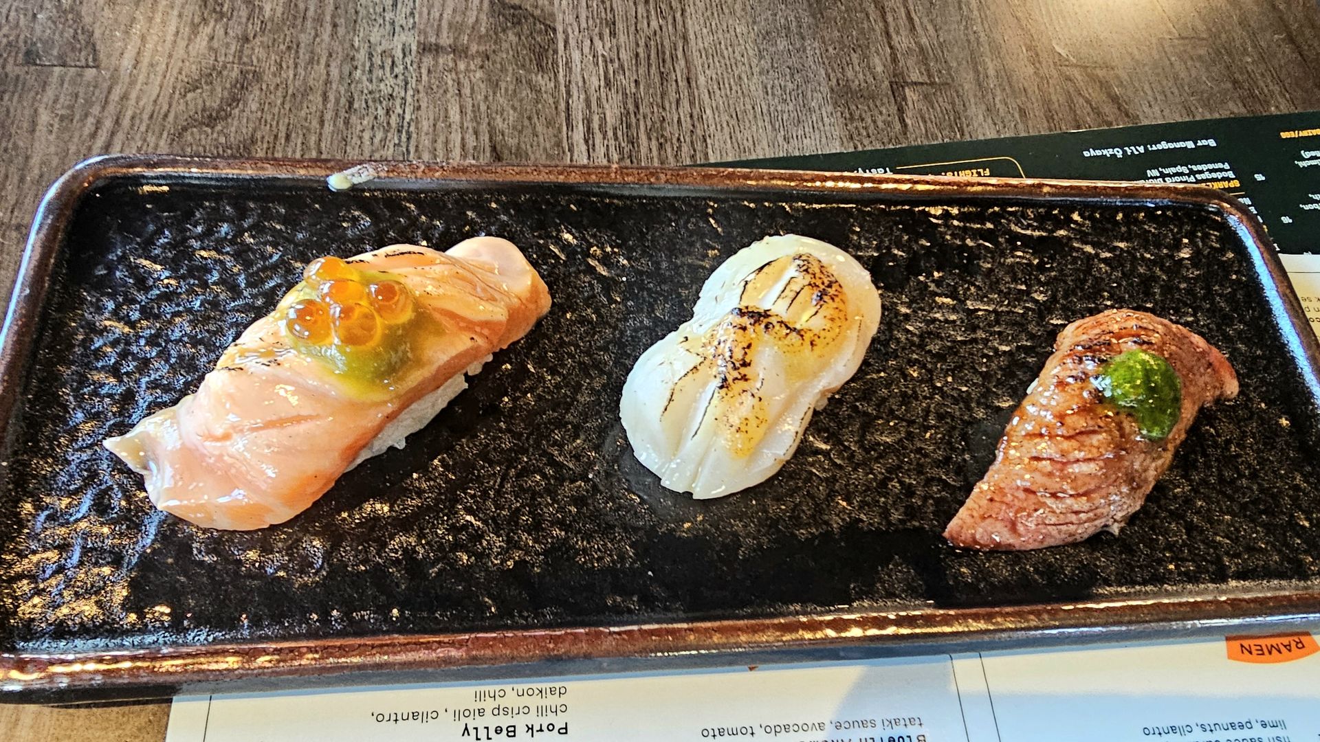 Assorted nigiri on a black tray: left salmon nigiri topped with bright orange salmon roe (ikura), center seared white fish nigiri with grill marks, right glazed pork belly nigiri with a green garnish.
