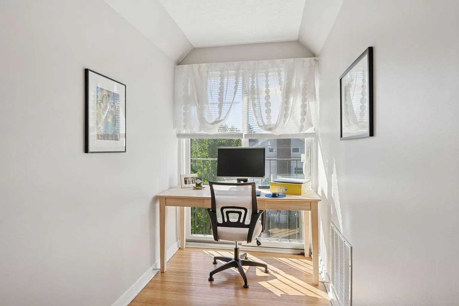 Bright, narrow office with wooden desk, black and white mesh chair, monitor, framed photos, and sheer white curtains covering large window with outdoor view.