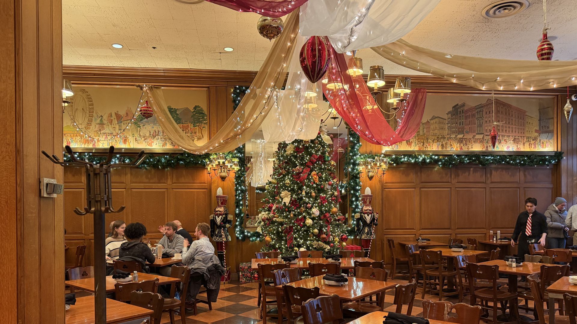 Interior of a restaurant decorated for Christmas with a large Christmas tree, nutcracker figures, hanging red and white drapes with lights, wood-paneled walls, and diners at tables.
