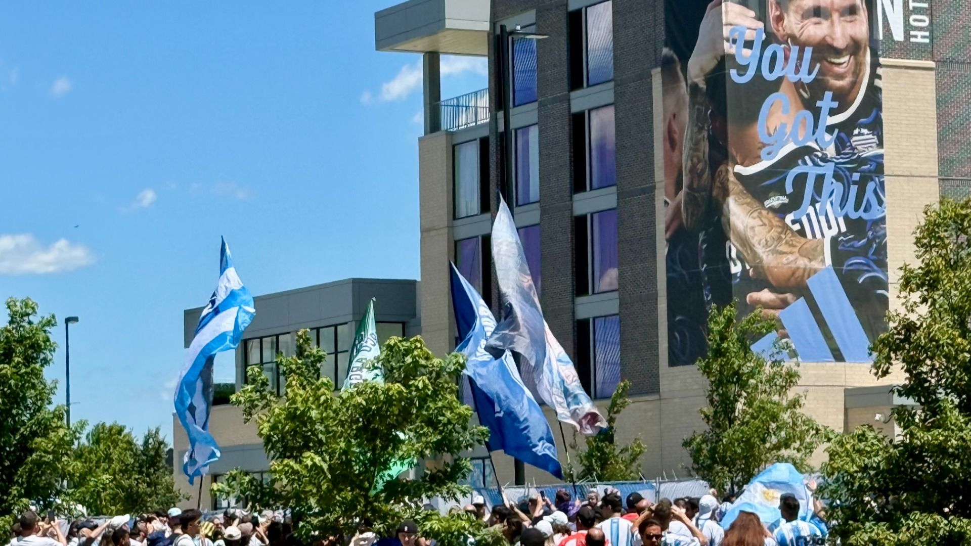 Bright day at a crowded outdoor event; a tall brick building features a large banner with a smiling man and the words "You Got This", while blue flags wave in front of trees and a crowd.