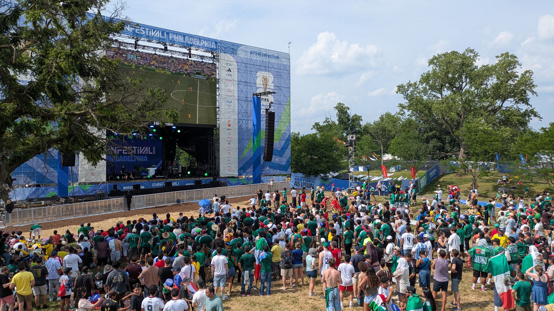 Outdoor FIFA Fan Festival stage with a giant screen showing a soccer match; a dense crowd in green and assorted colors watches from a park as trees line the scene under a blue sky with banners.