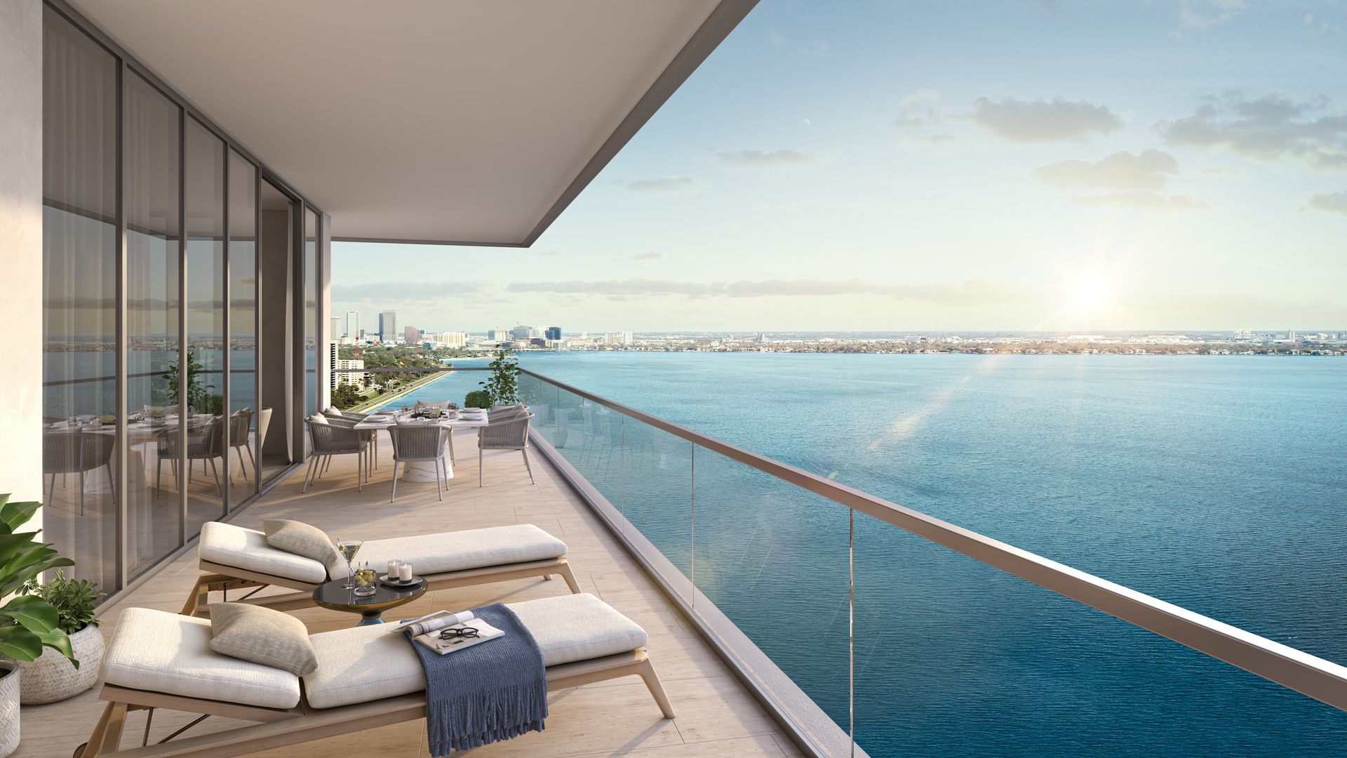 A patio with two chaise lounges with cream cushions and a dining table overlooking cerulean water and a few high-rise buildings 