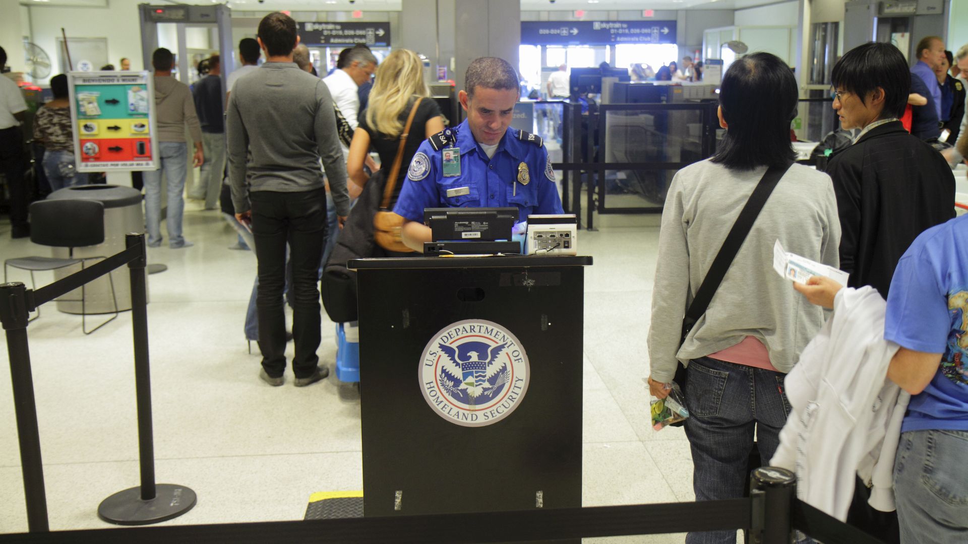 In this image, a man sits behind a DHS podium in an airport security line. A line of people stand next to him.