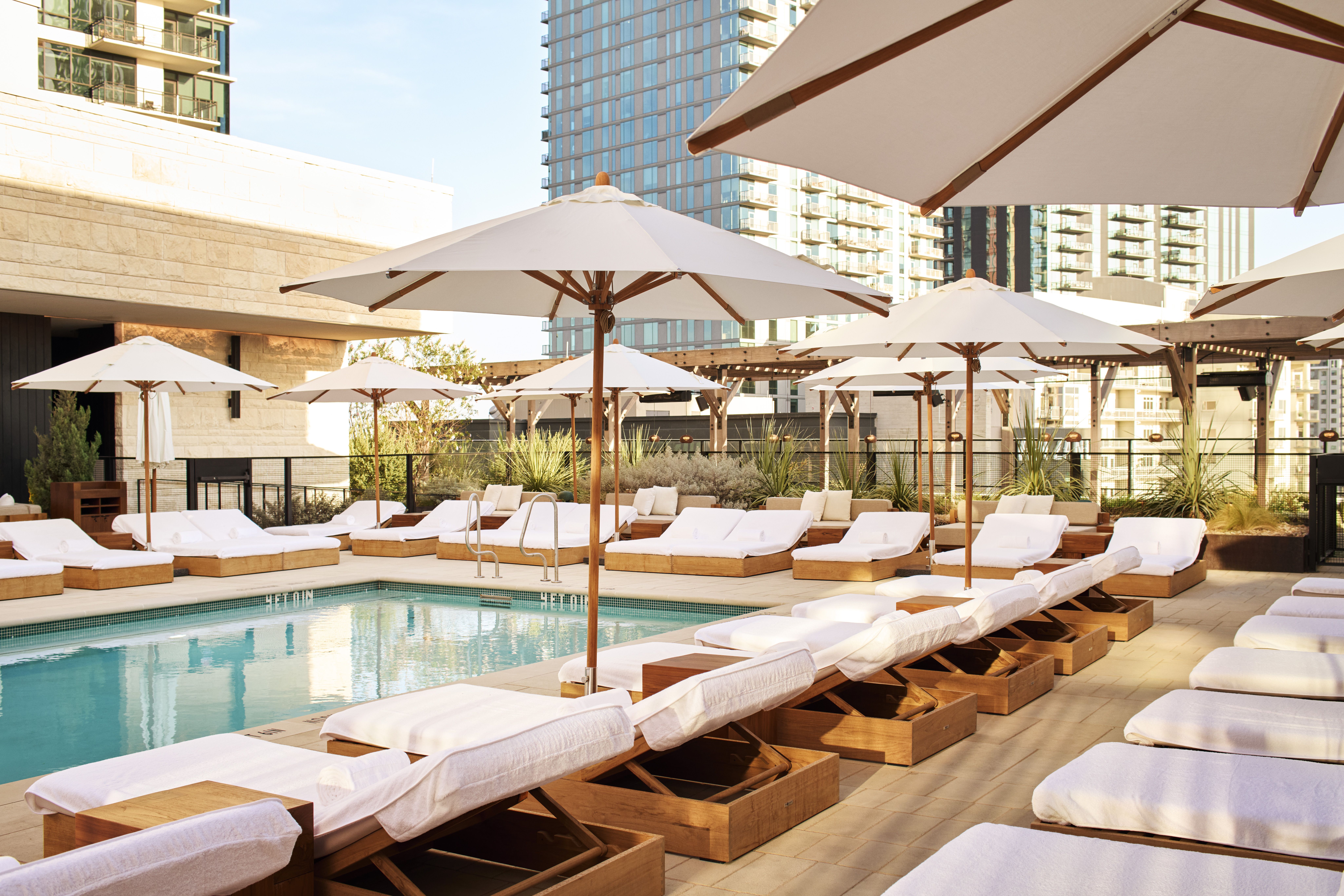 Poolside rooftop scene with wooden sun loungers and white cushions, large white umbrellas, turquoise pool in foreground, and tall modern buildings in the background.