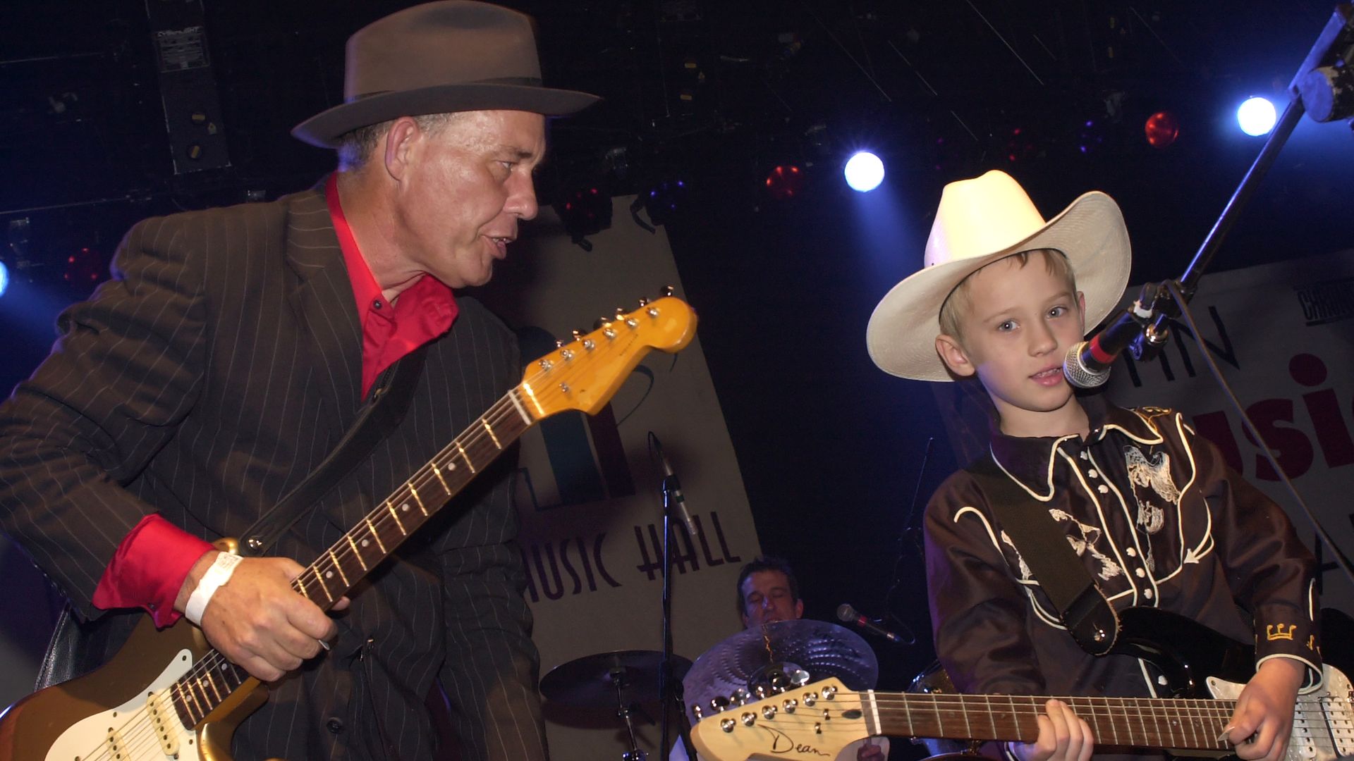 Two musicians on stage: an older man in a pinstripe suit and brown hat plays a sunburst electric guitar, while a young boy in a white cowboy hat sings into a microphone.