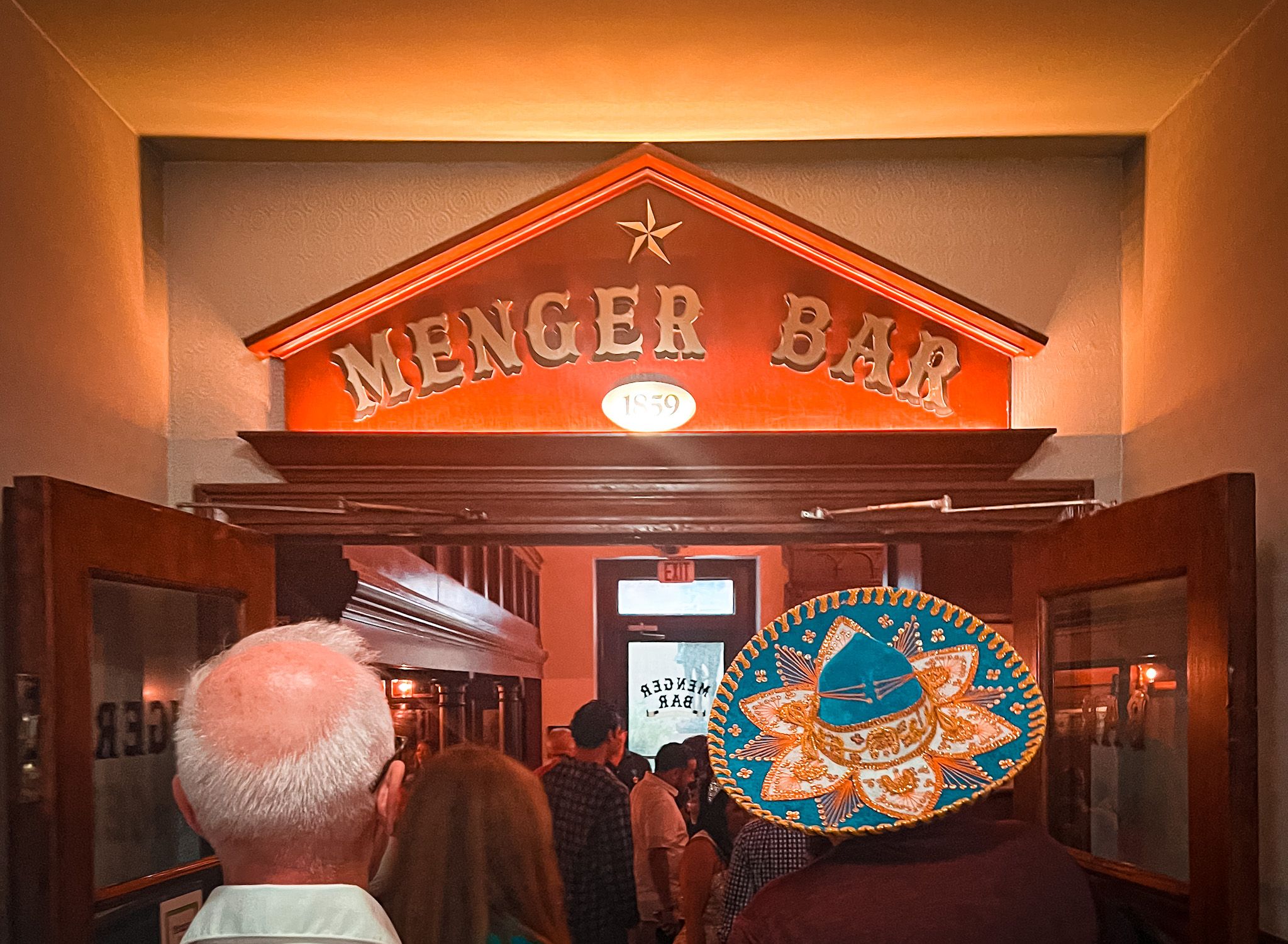 A group of people pass through a door with the words "Menger Bar" overhead. 
