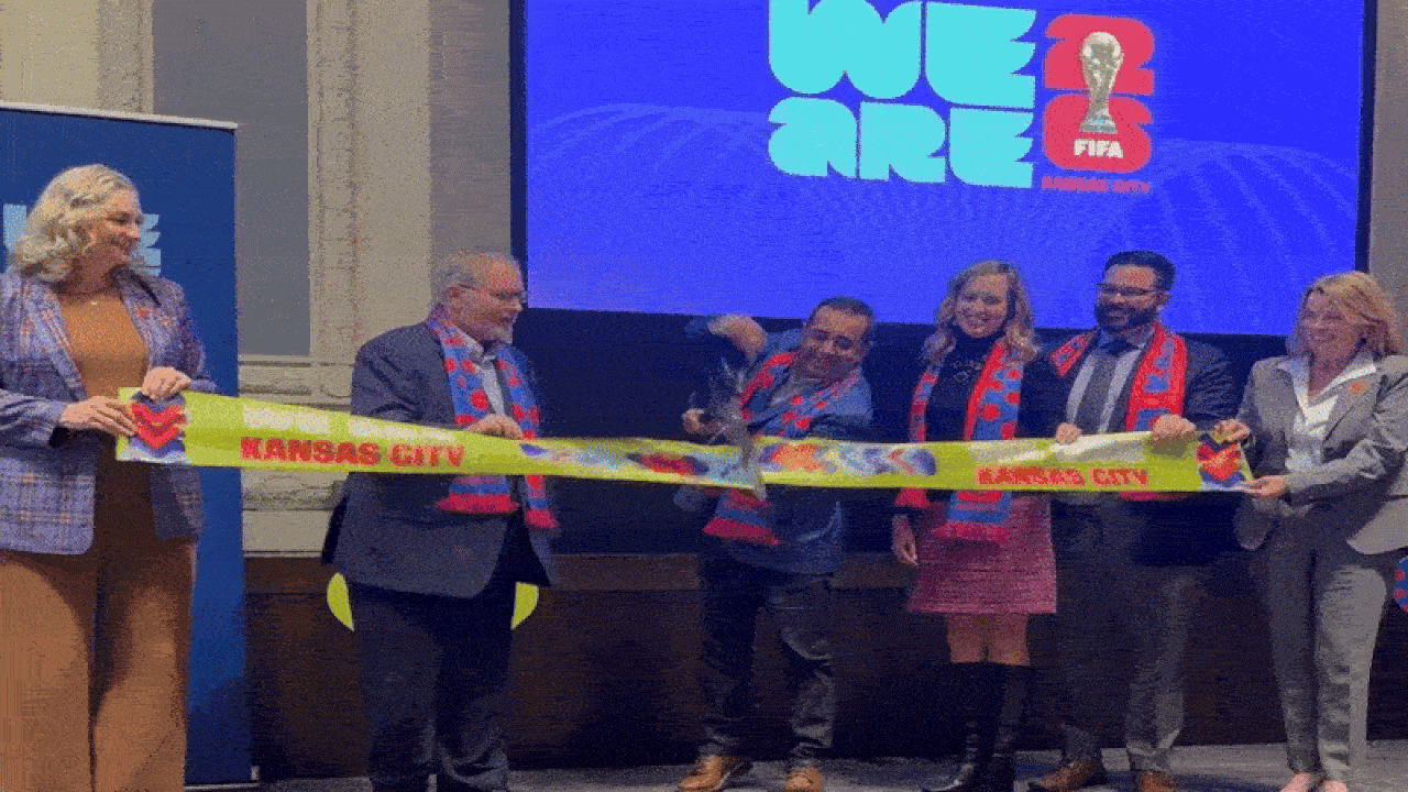 Six people cut a bright yellow ribbon reading "Kansas City" at a ceremony, wearing blue and red scarves, with a blue screen behind them showing "We Are FIFA" and a trophy logo.