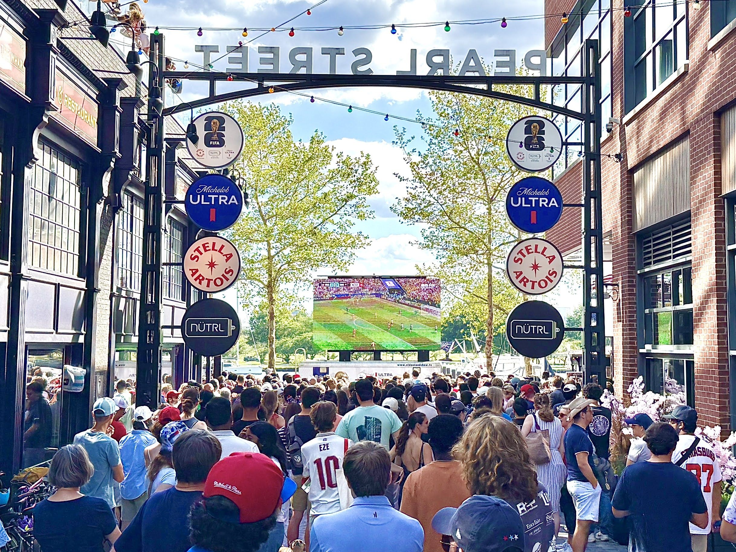 Crowded urban festival alley with brick buildings, string lights, and circular beer signs (Michelob Ultra, Stella Artois). A big screen in the distance shows a soccer game; people seen from behind.
