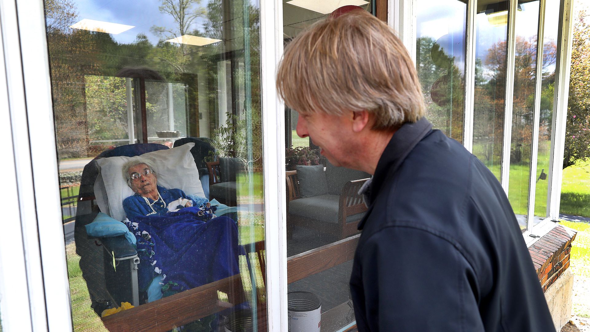 A man visiting his mother through the window of a nursing home.