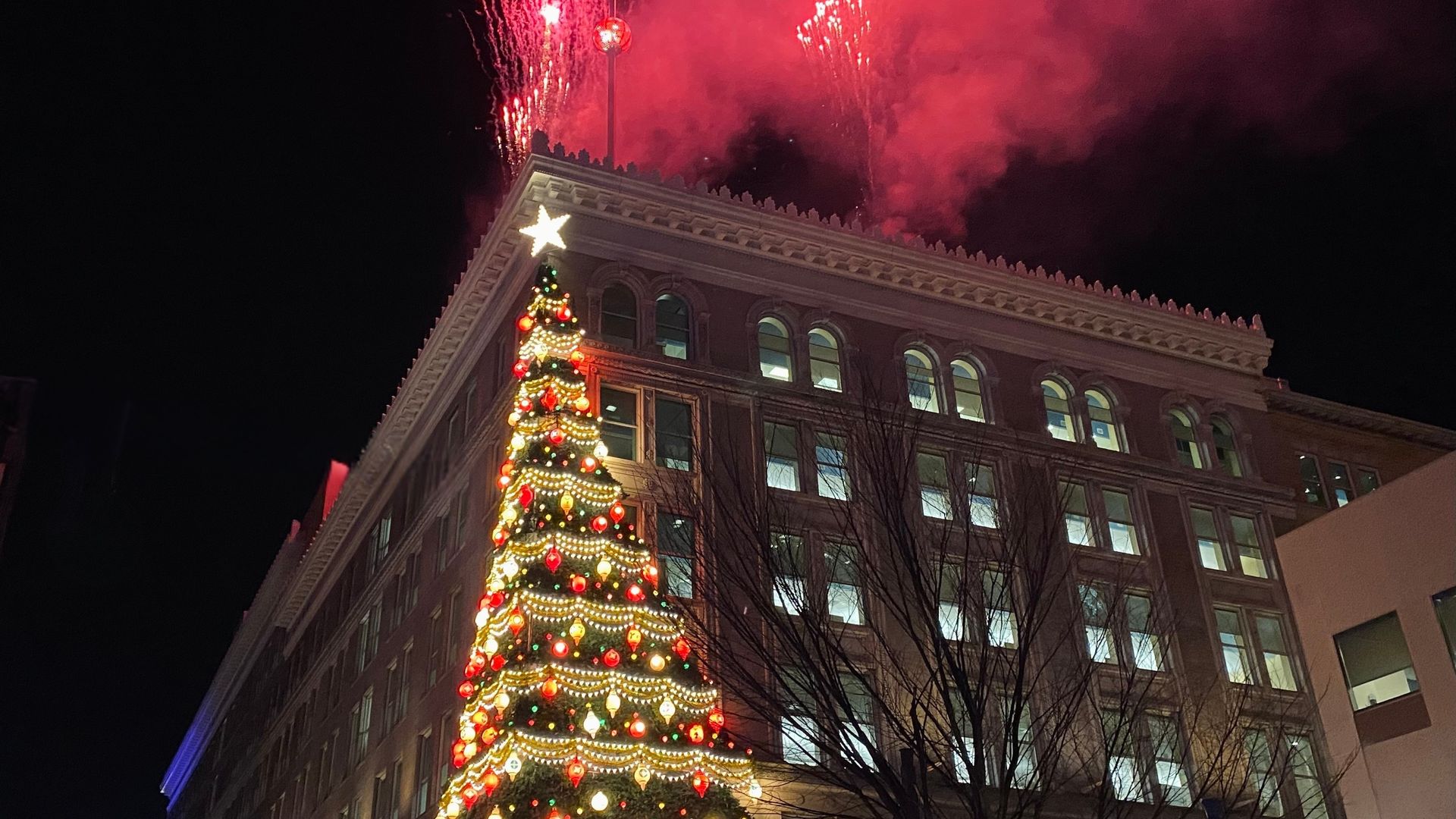 Night scene of a large decorated Christmas tree with red, gold lights and a glowing star in front of a multi-story building, with red fireworks and smoke in the sky above.