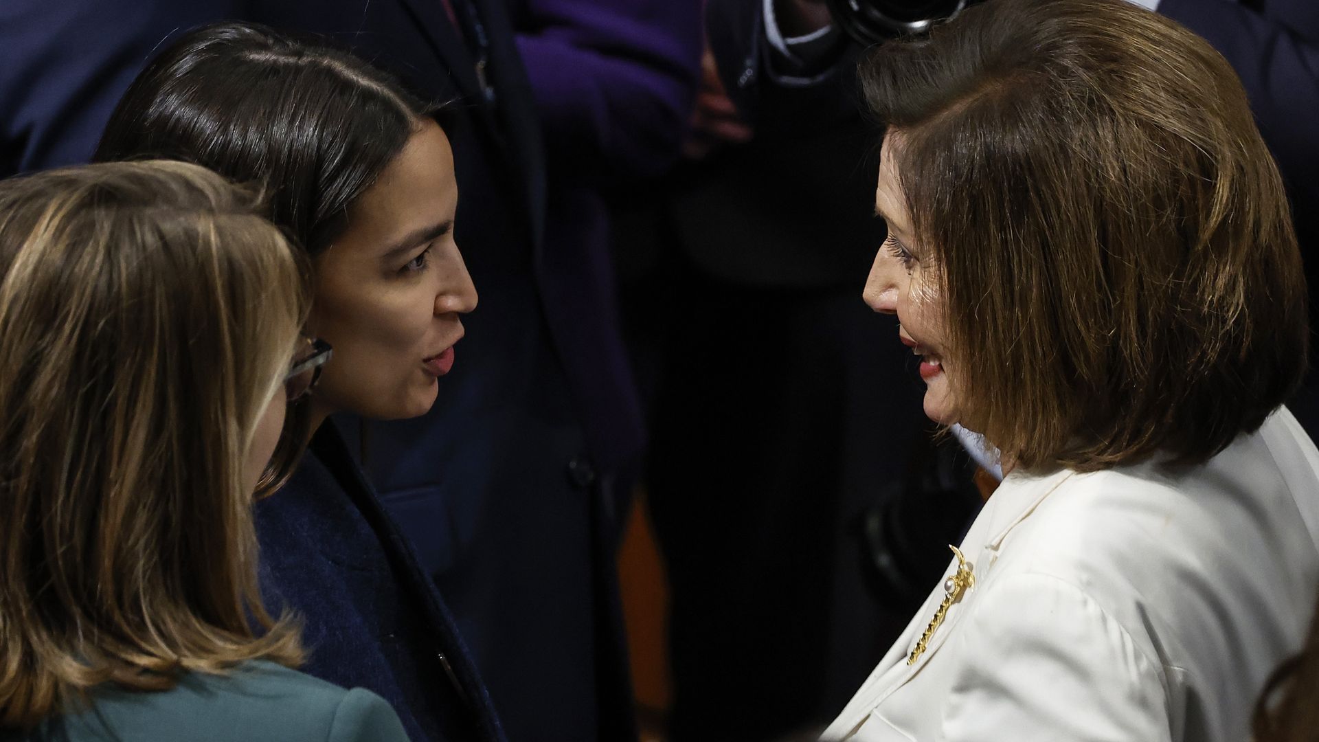 Two women facing each other in a close conversation; left woman with dark hair and teal top, right woman wearing a white blazer with a gold chain, against a dark background.
