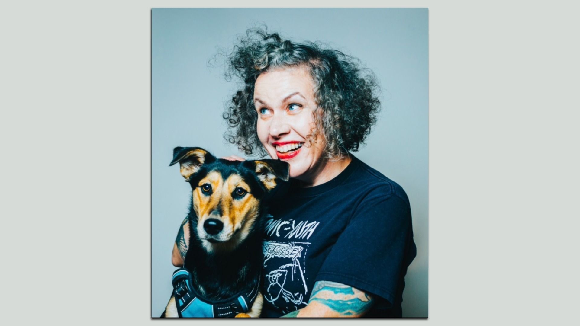 Smiling person with curly gray hair and red lipstick holds a brown-and-black dog with a blue harness; pale blue backdrop.