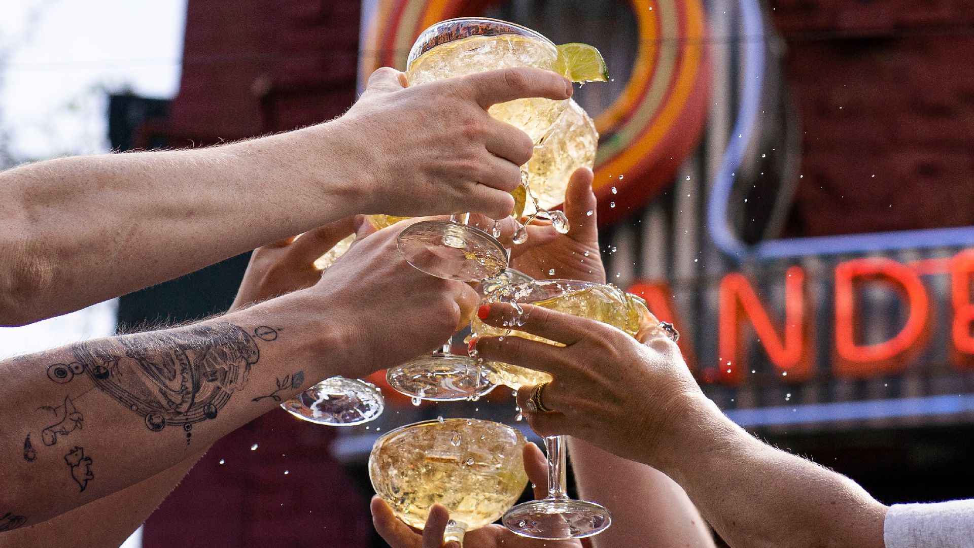 Several hands clink crystal champagne glasses in a festive toast; lime wedges on rims, champagne splashes, visible tattoos on a forearm, and a colorful neon sign in the blurred background.