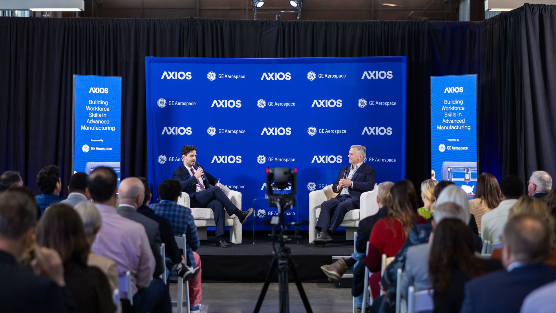 Two men in suits seated on stage in front of a blue backdrop with Axios and GE Aerospace logos, speaking to an audience at an event titled Building Workforce Skills in Advanced Manufacturing.