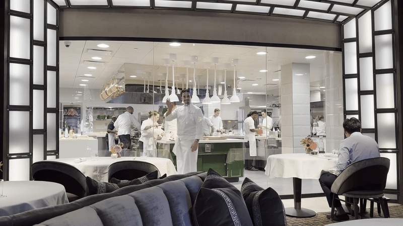 A man in a chef's coat waves from behind a glass wall.