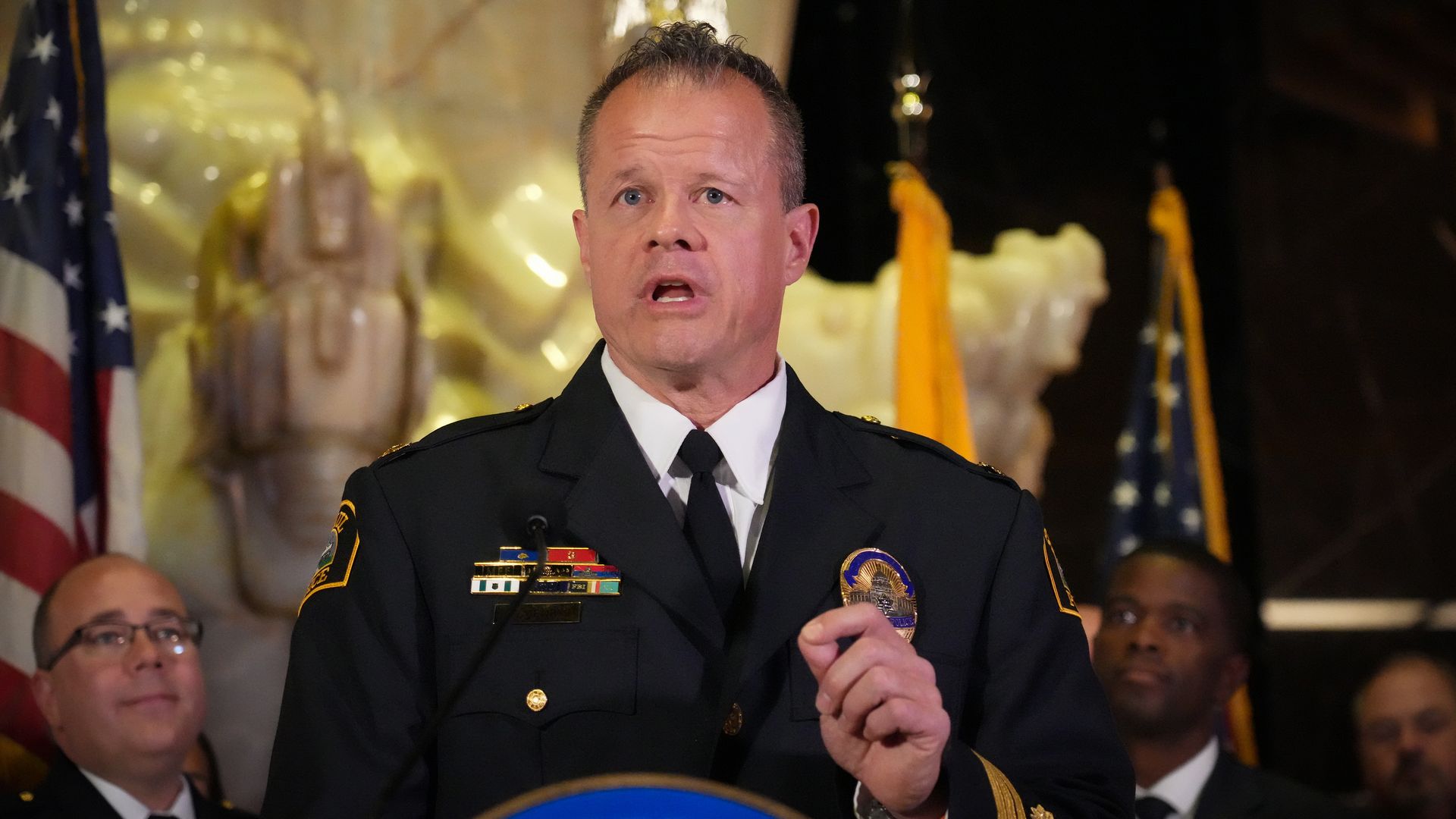 A uniformed police officer speaking at a podium with a blue emblem, flanked by American flags and other officials in the background, inside a decorated building.