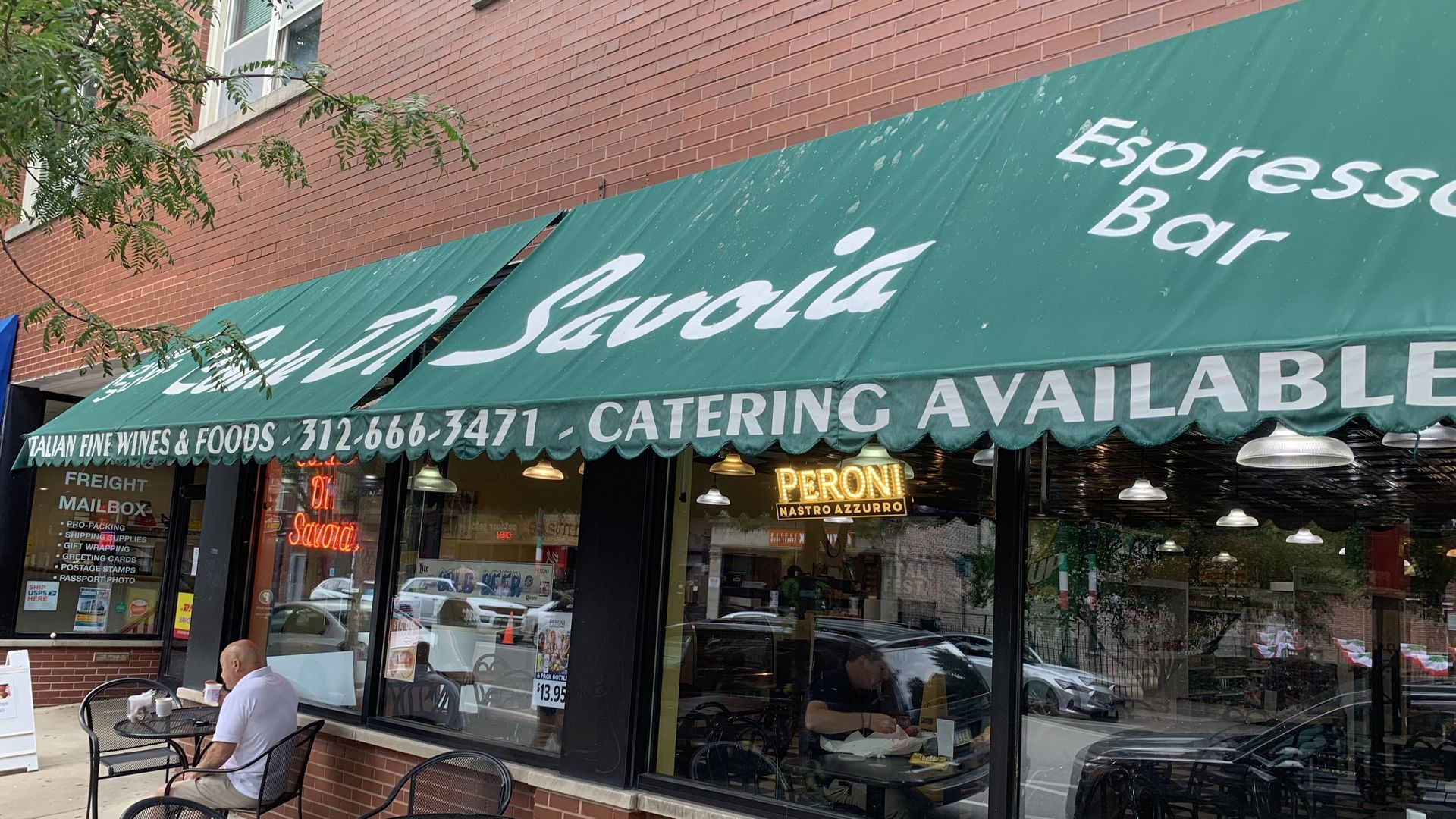 Brick storefront with a large green awning and outdoor cafe seating; a man sits at a table while tree branches frame the left. Signs read "Italian Fine Wines & Foods" and "Catering Available."