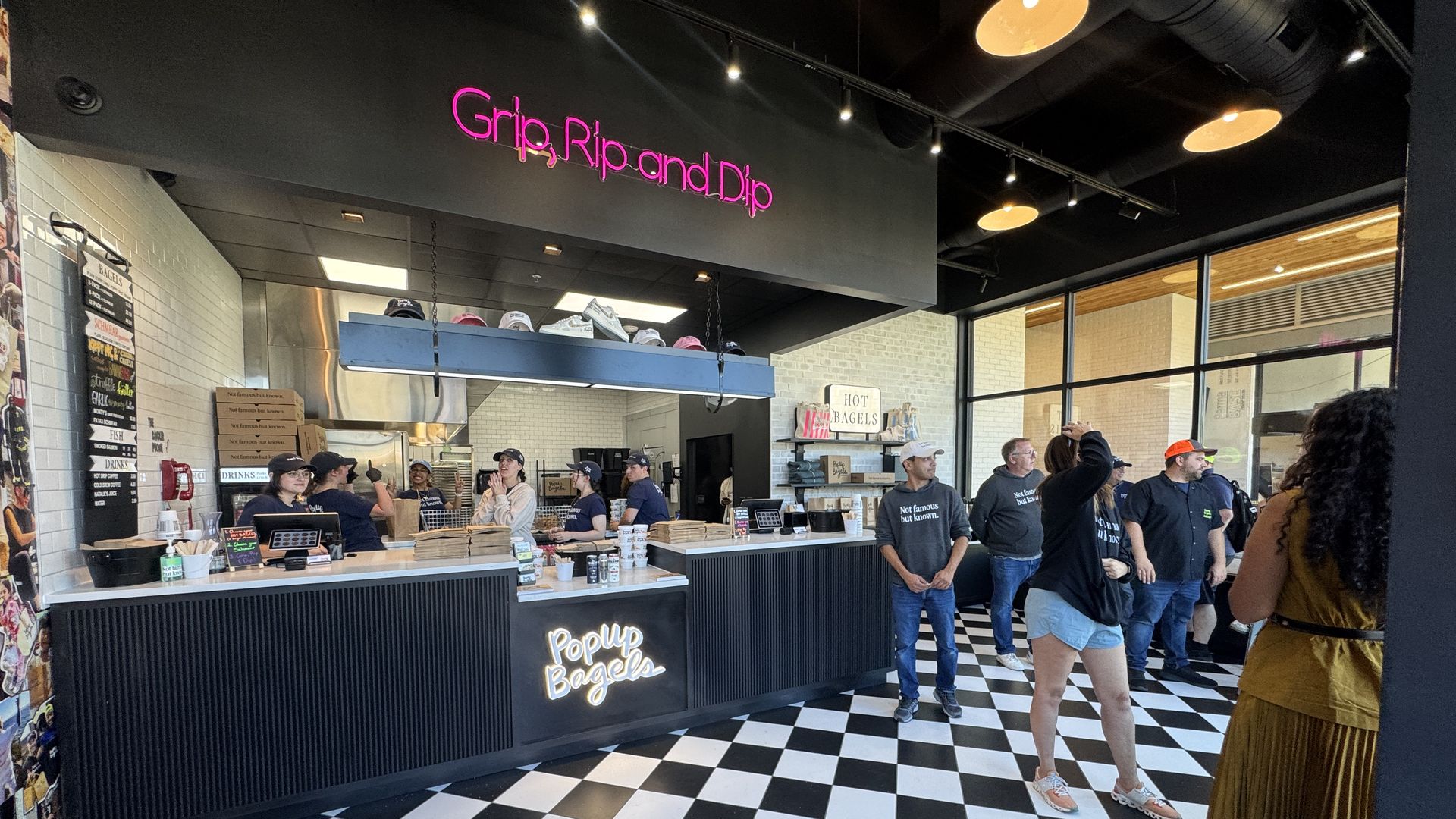 Interior of a bagel shop with black and white checkered floor. Staff wearing black shirts and caps behind counters. Neon sign reads Grip rip and dip