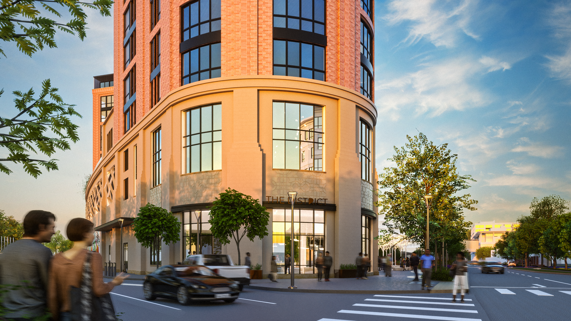 Street view of a red brick and beige modern building with large windows, labeled "THE DISTRICT," surrounded by trees, pedestrians, and cars under a blue sky with clouds.