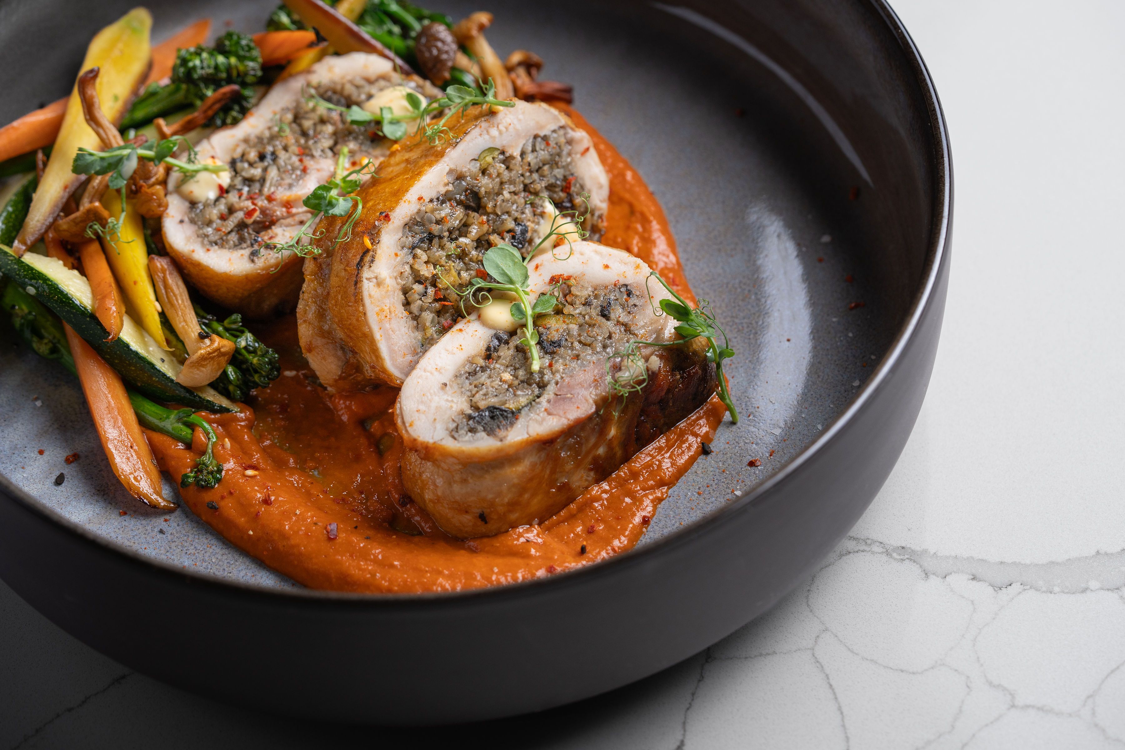 Three slices of stuffed chicken breast on red sauce, garnished with microgreens, served with sautéed mixed vegetables including carrots, zucchini, and mushrooms in a black bowl.