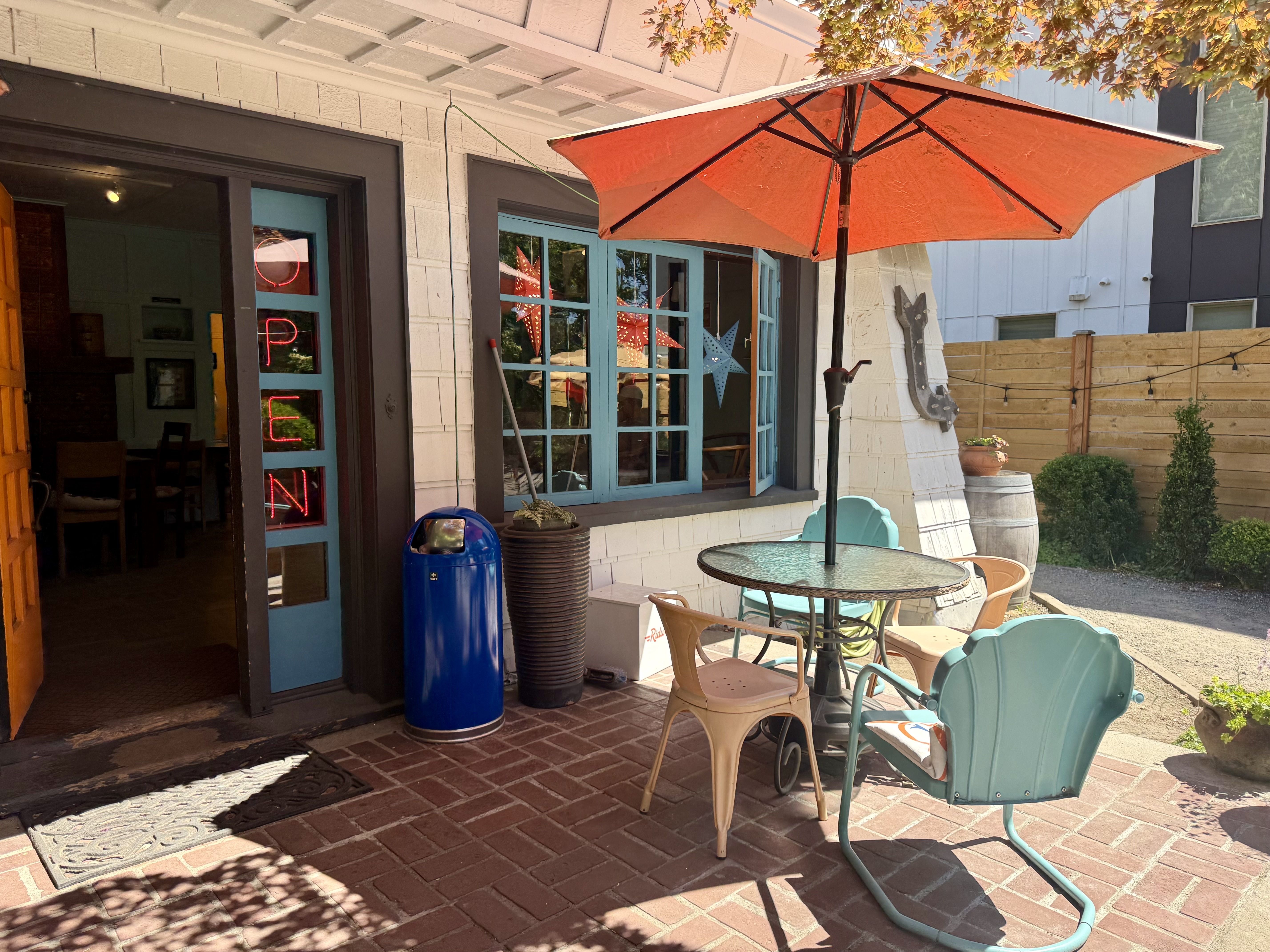 Sunlit outdoor cafe patio with a large orange umbrella over a glass table, turquoise doors and windows, pastel chairs, and a red neon OPEN sign in the doorway.