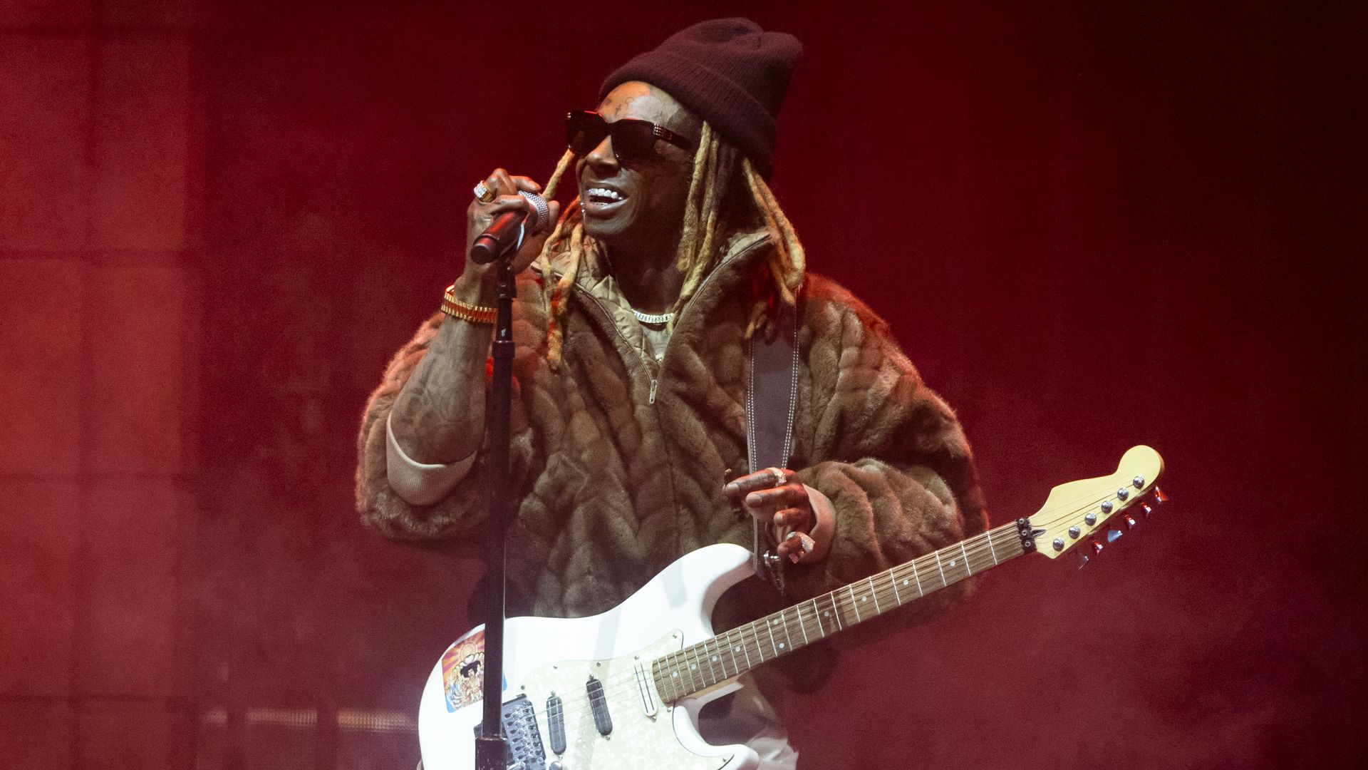 Musician wearing a brown fur coat, red beanie, and sunglasses performs on stage with a white electric guitar and microphone against a red smoky background.
